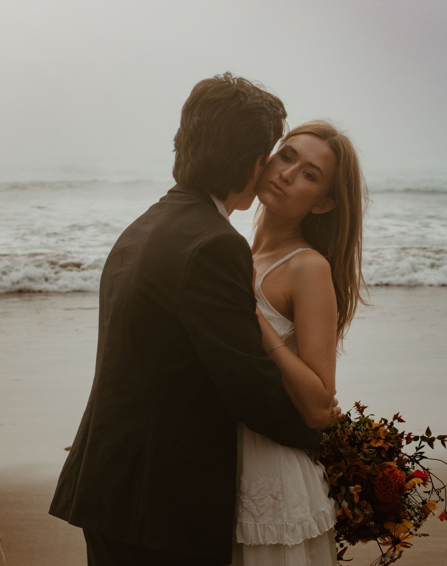Couple in wedding attire share an intimate moment holding a bouquet by the ocean.