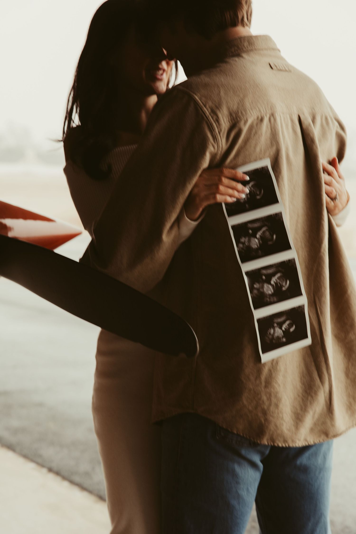 Couple embracing near a red plane while woman holds ultrasound photos against his back in a hangar.