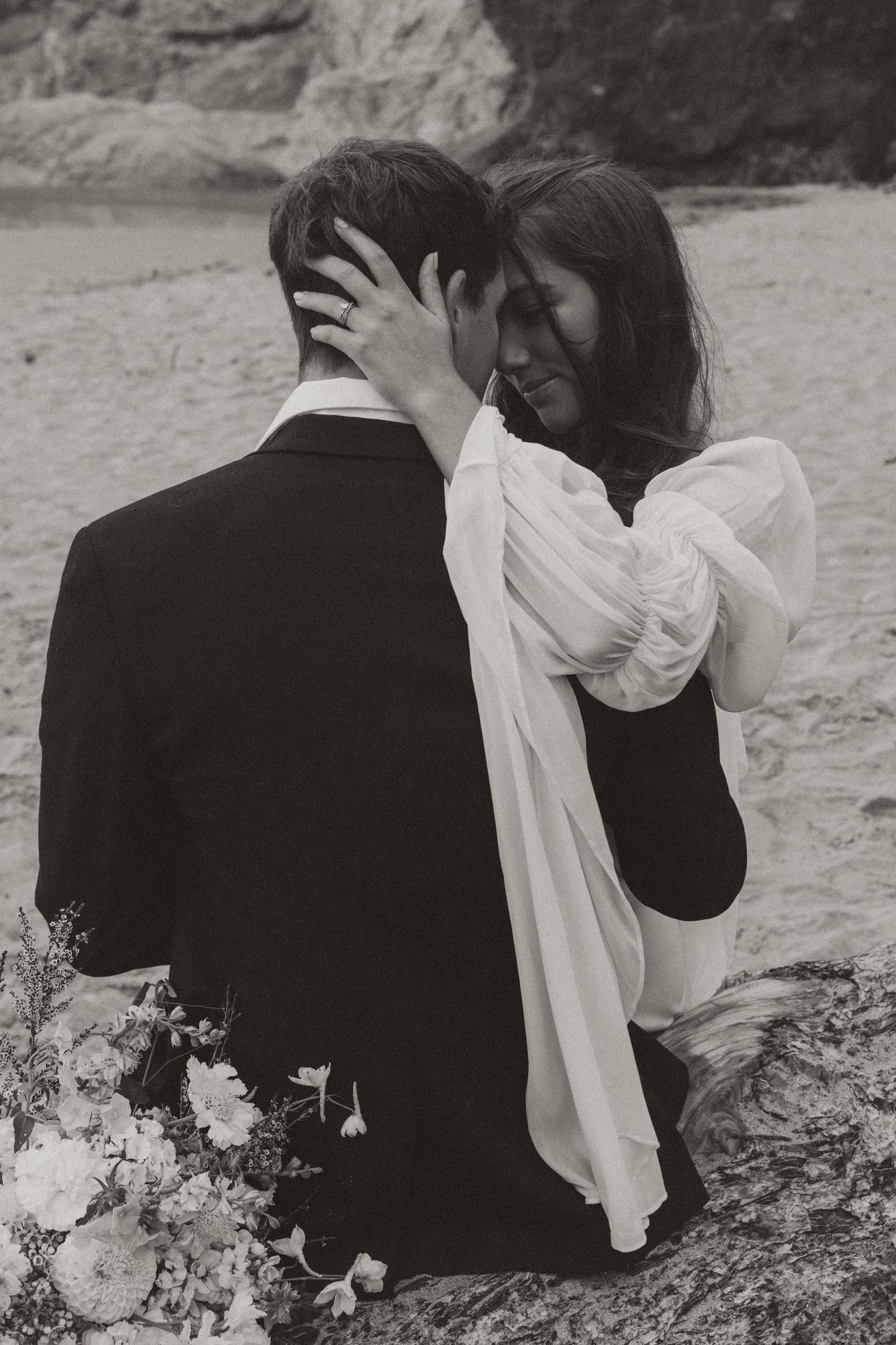 Black and white photo of bride embracing groom from behind on a rocky beach.