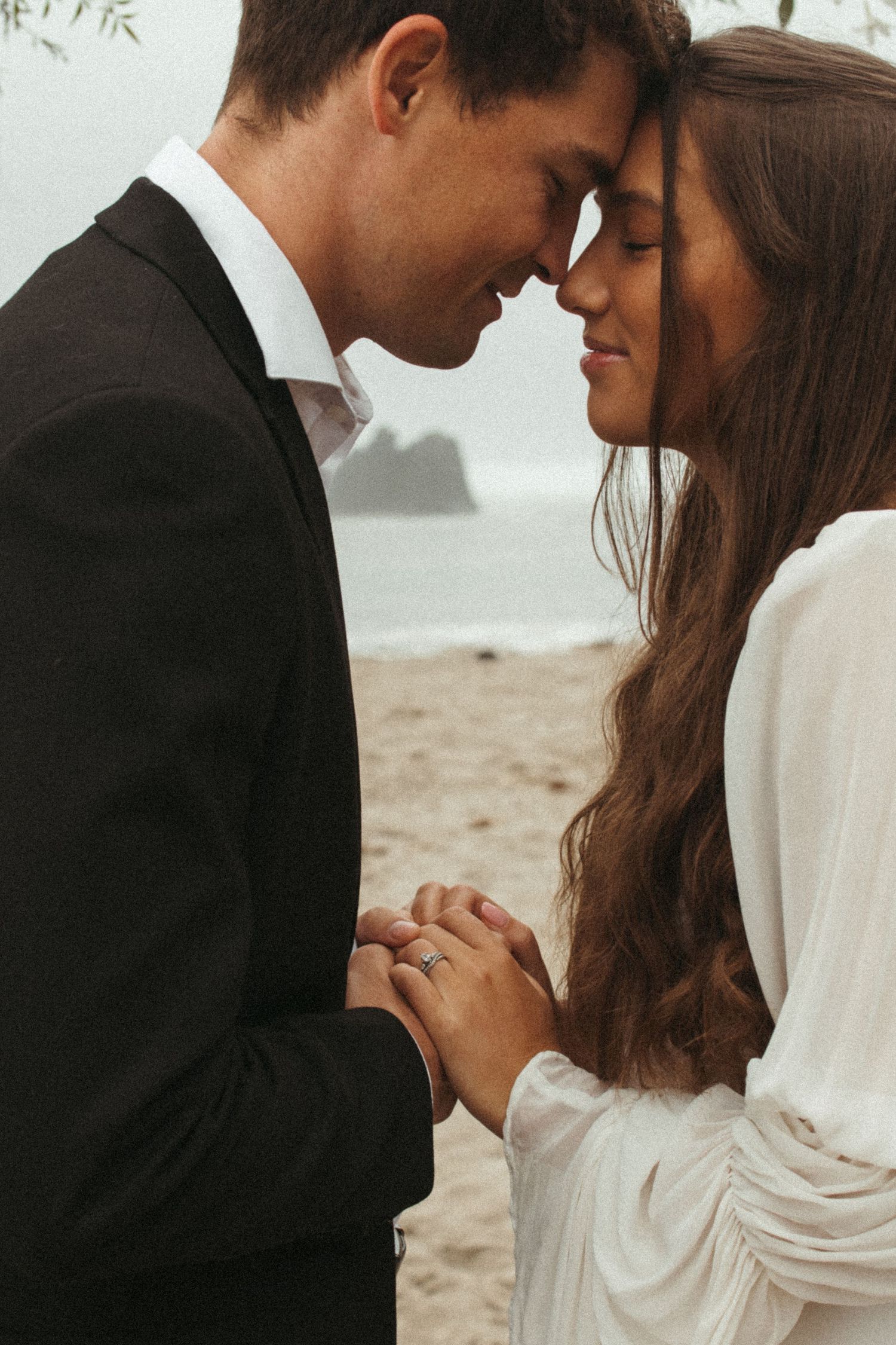 Bride and groom touch foreheads tenderly while holding hands on a foggy beach.