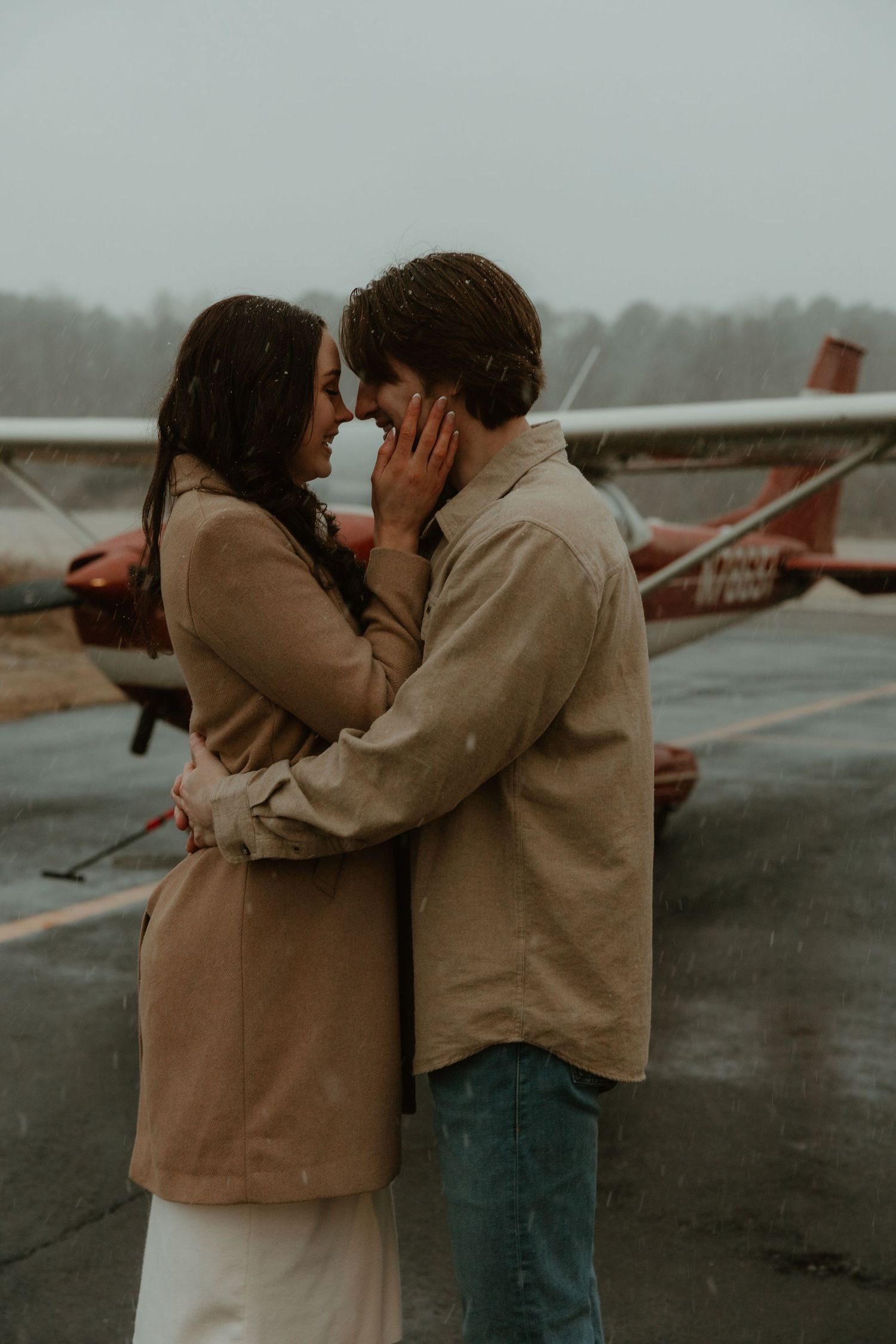 Couple embracing in the rain beside a vintage red airplane during an outdoor engagement session.