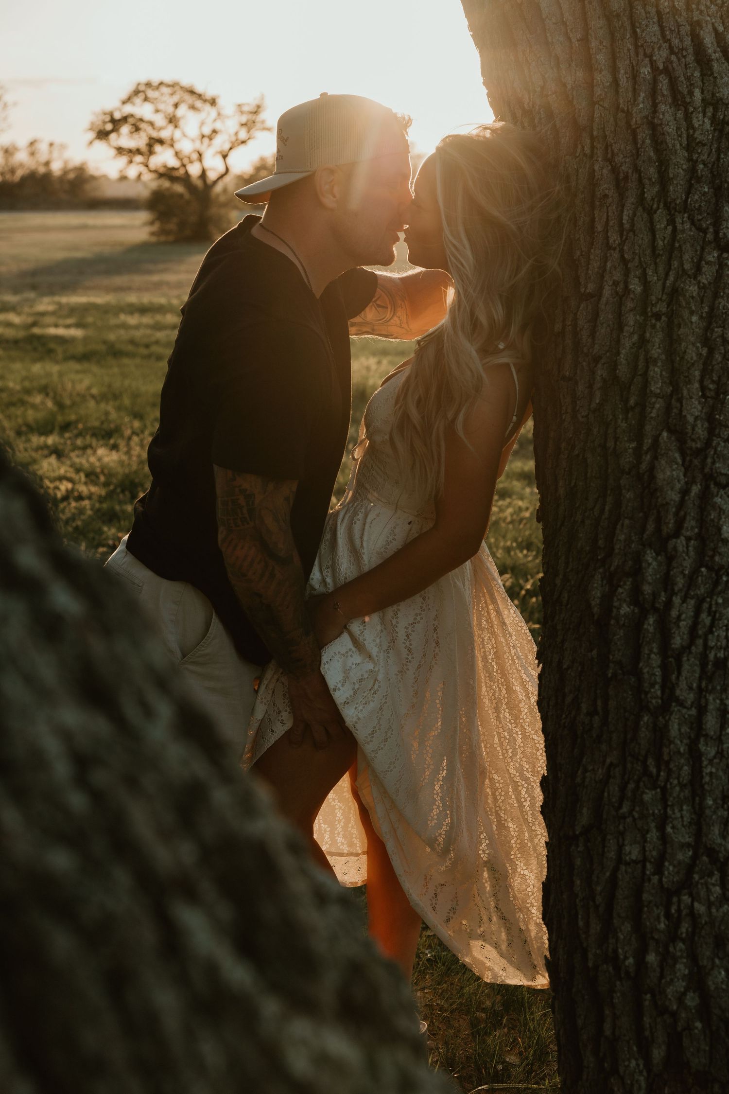 Couple sharing a kiss at golden sunset beside a large tree in an open countryside field.