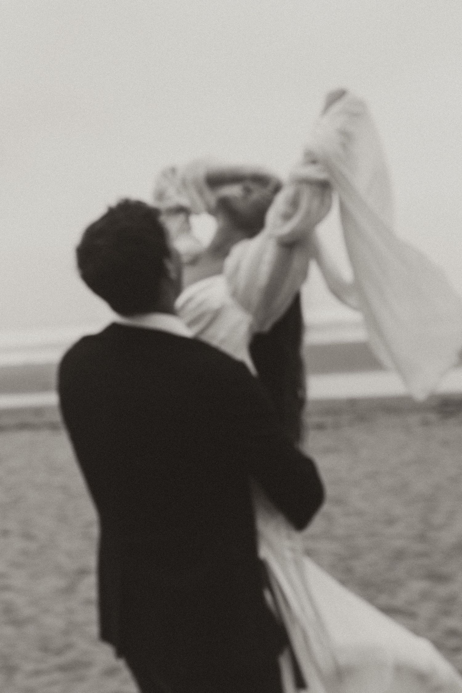 Black and white photo of groom lifting bride on a sandy beach in a romantic wedding moment.