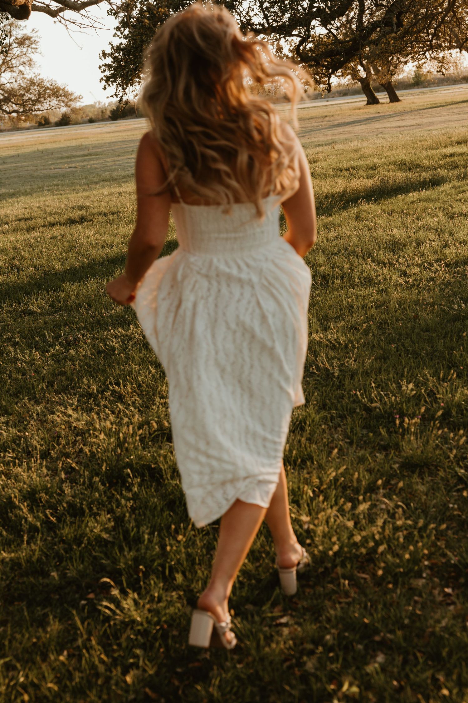 Blonde woman in white lace midi dress and heels walking away through a sunlit green park at sunset.