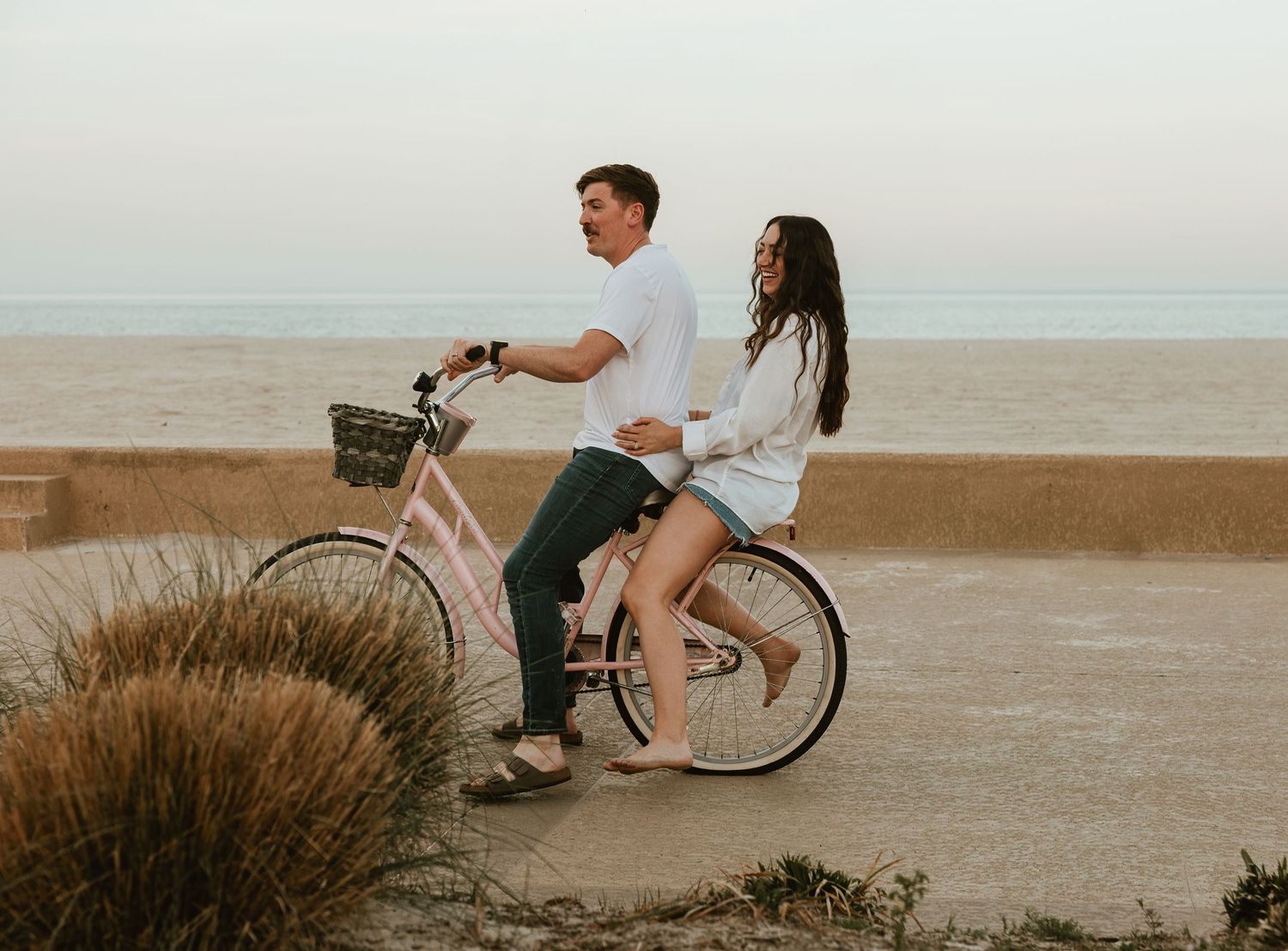 Romantic couple riding a pink beach cruiser bicycle together along a scenic ocean overlook.
