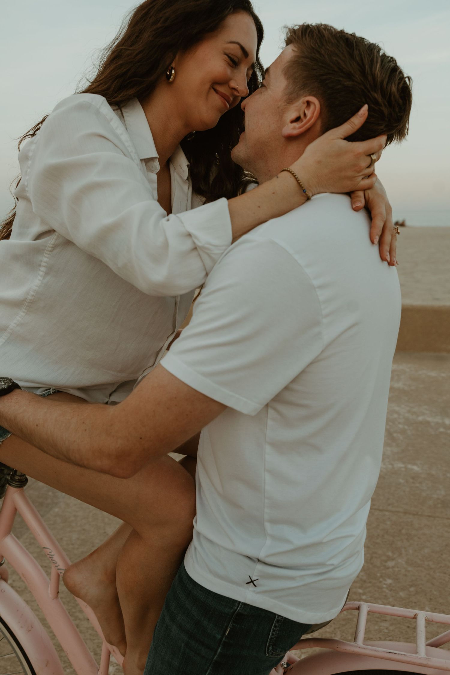 Woman sitting on pink bicycle leaning in to kiss a man in white t-shirt near a sandy beach.