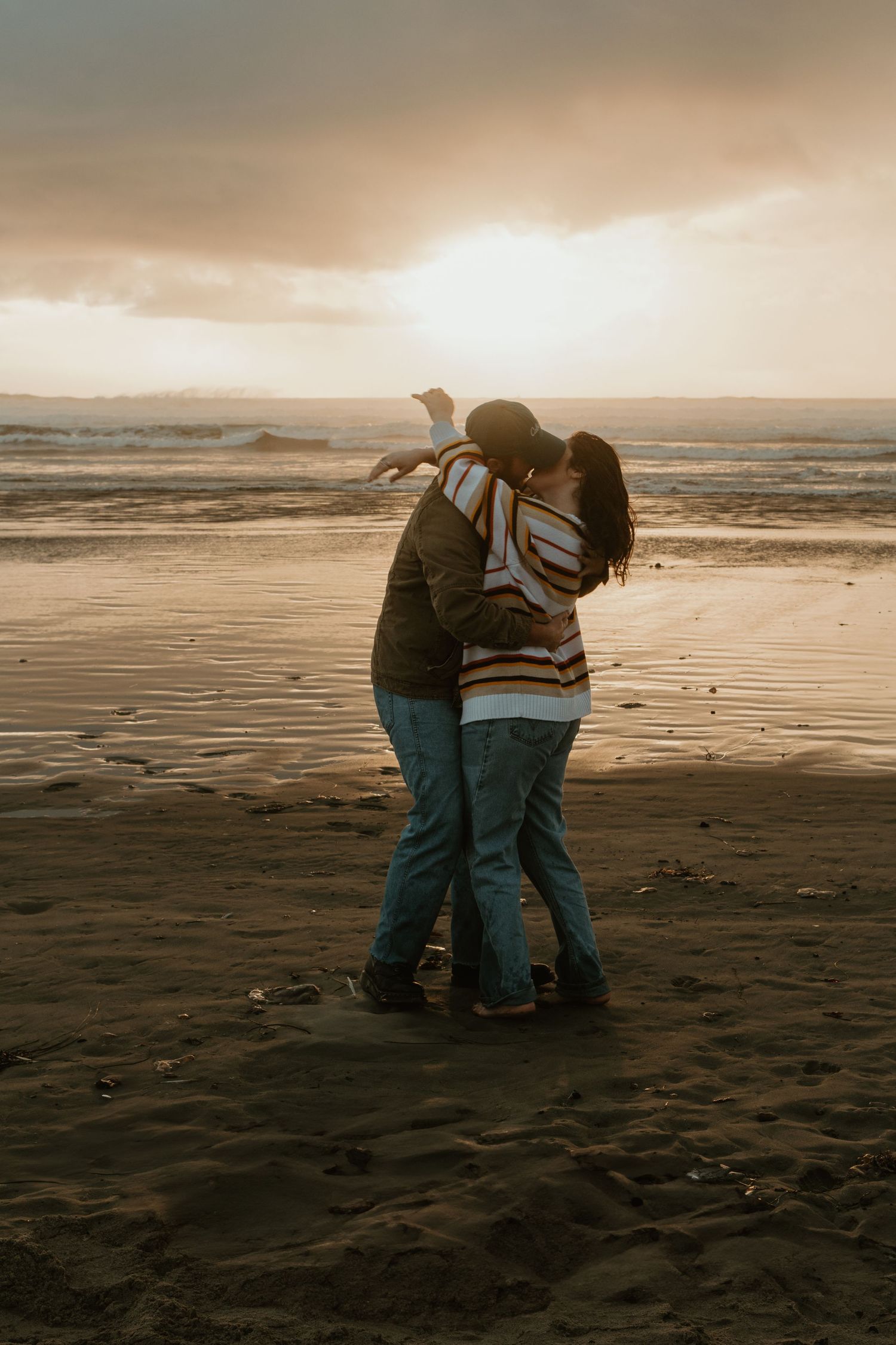 Couple sharing a romantic kiss on a sandy beach during a dramatic golden sunset.