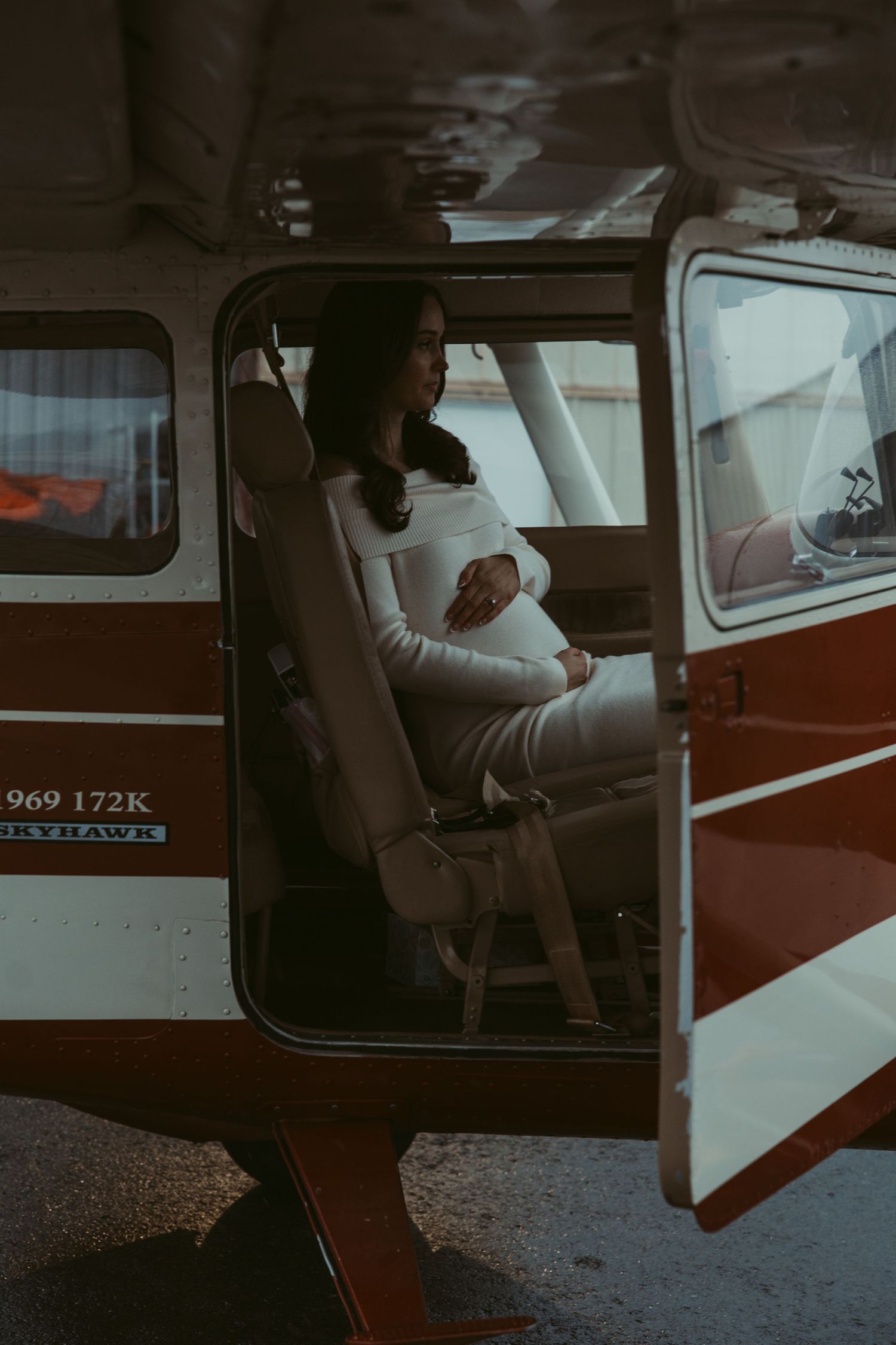 Pregnant woman in white outfit sits gracefully in open door of a vintage red and white plane.