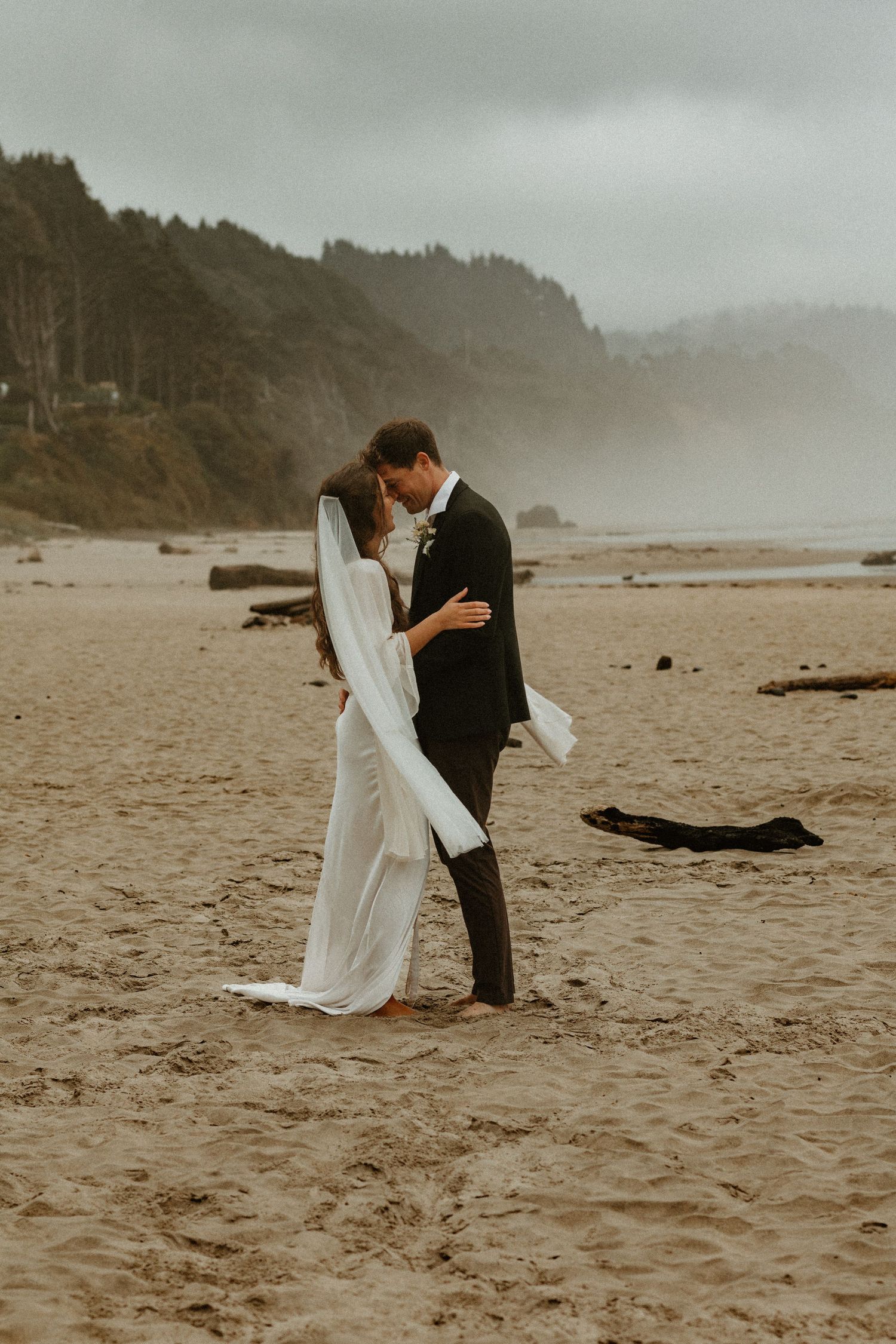 Bride and groom share a romantic embrace on a misty, fog-covered sandy beach.