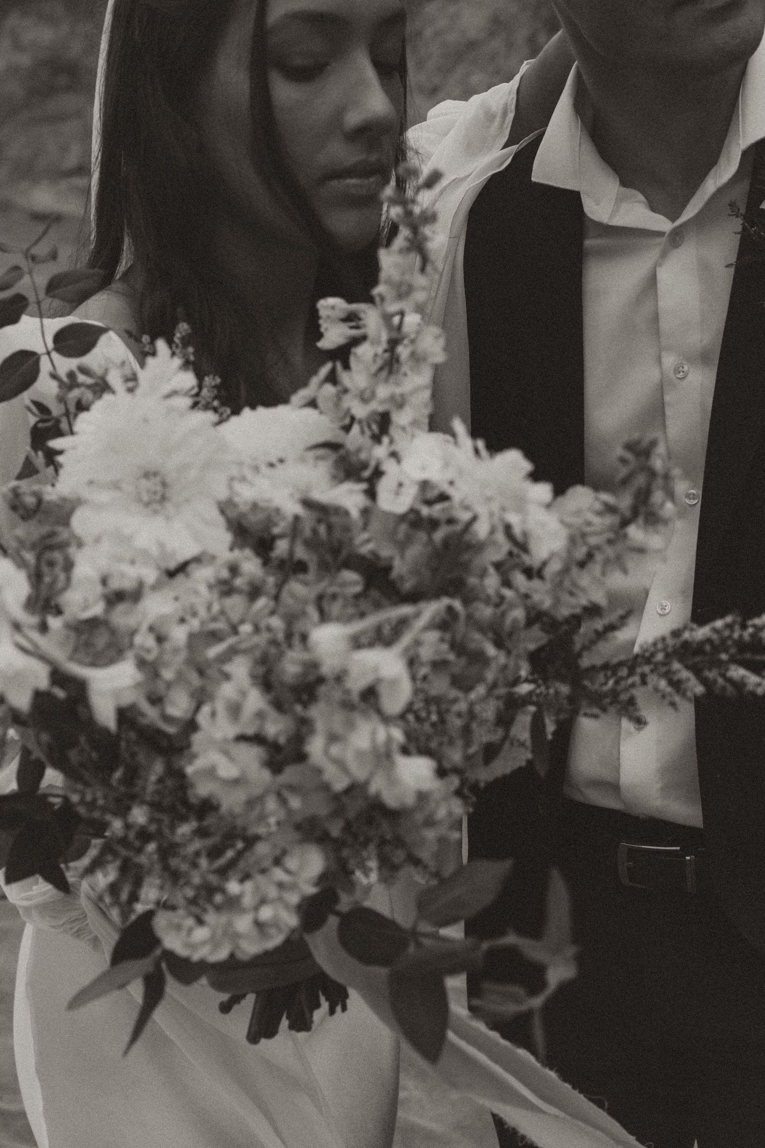Black and white close-up of bride and groom holding a lush floral wedding bouquet with dahlias and wildflowers.