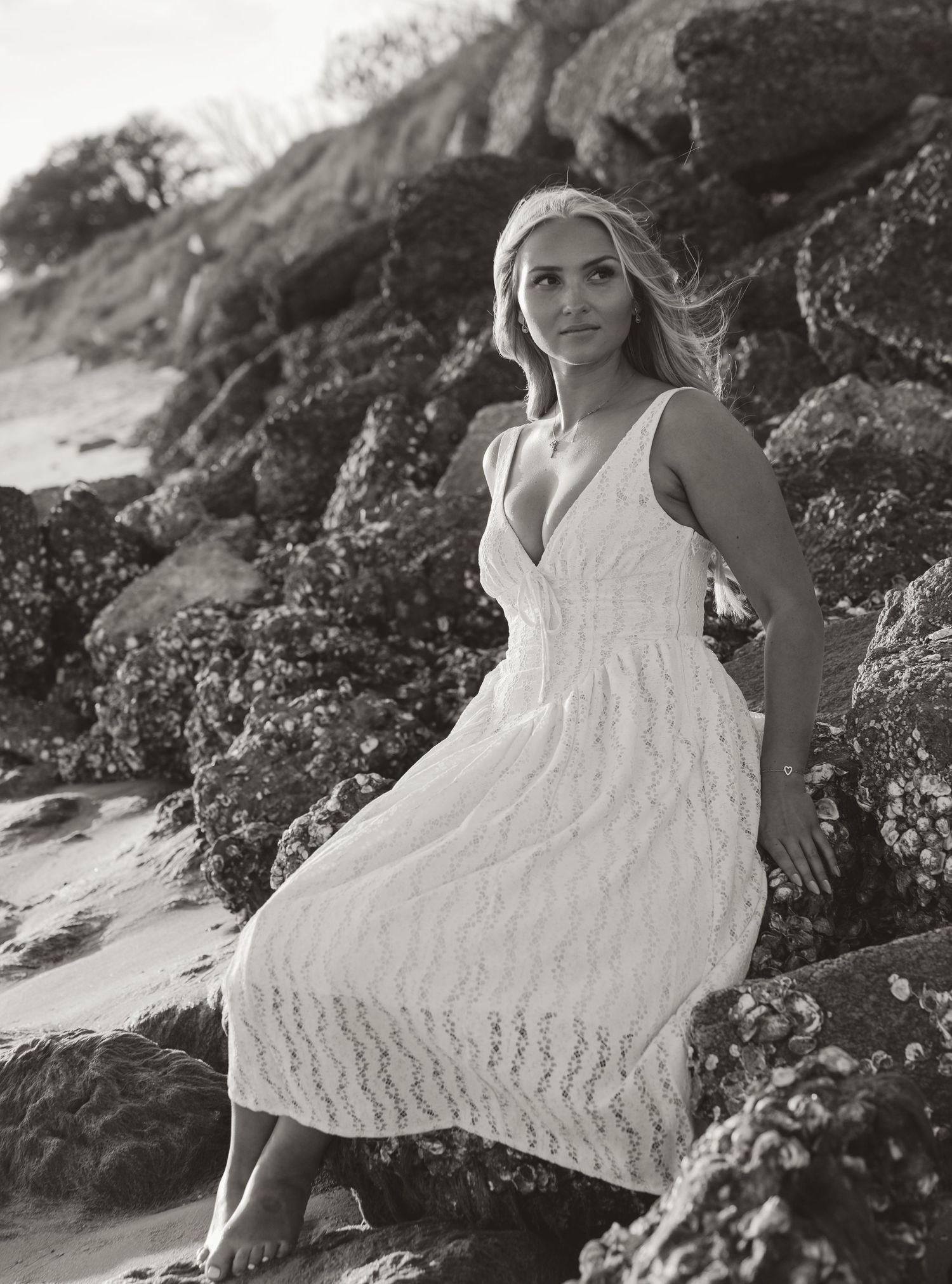 Black and white portrait of a blonde woman in a white lace midi dress sitting on rocky coastal boulders.