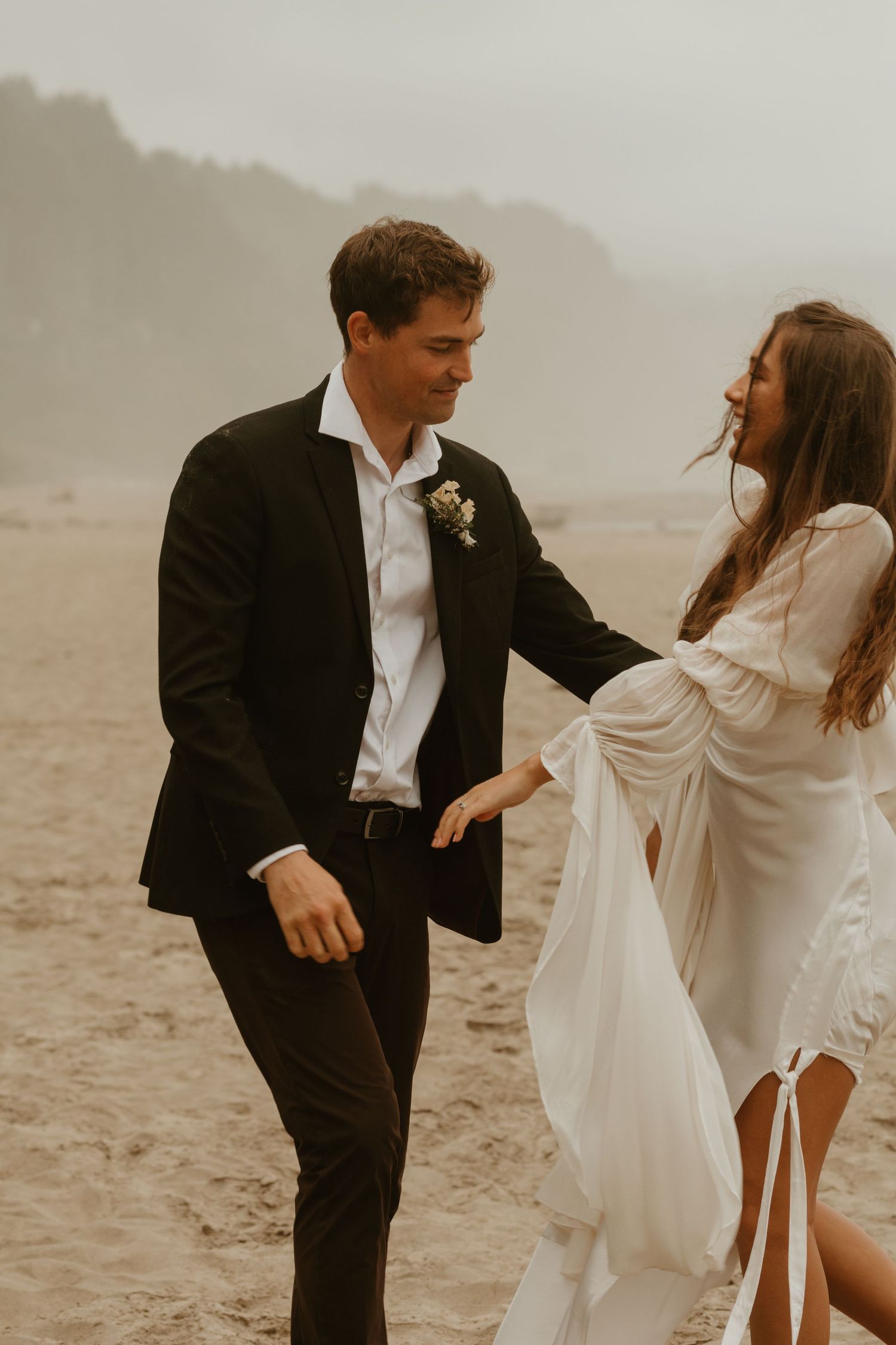 Bride and groom walking on a misty beach, bride in flowing white dress holding her skirt while groom wears black suit.