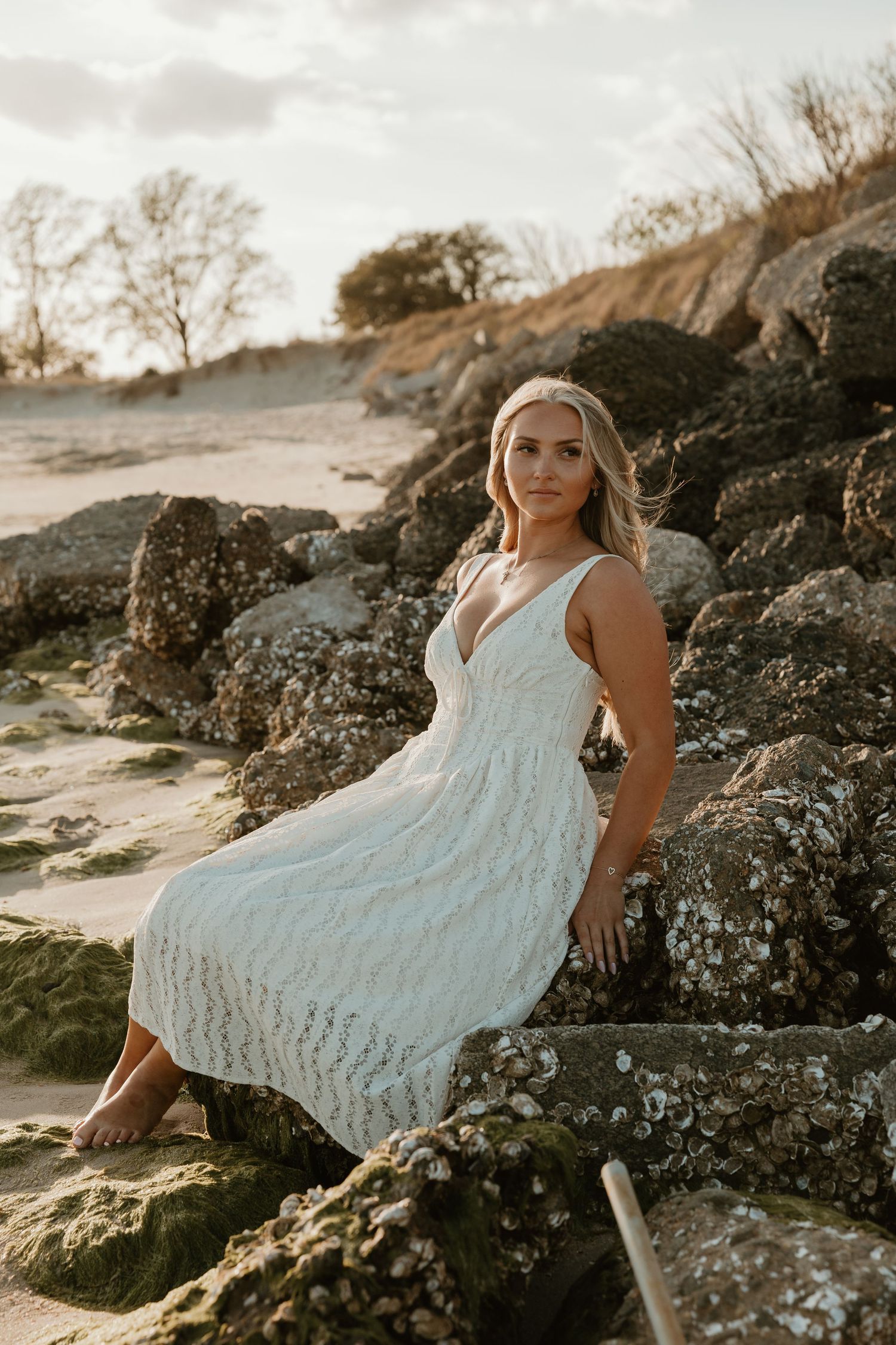 Blonde woman in white lace dress sitting on rocky beach at sunset.