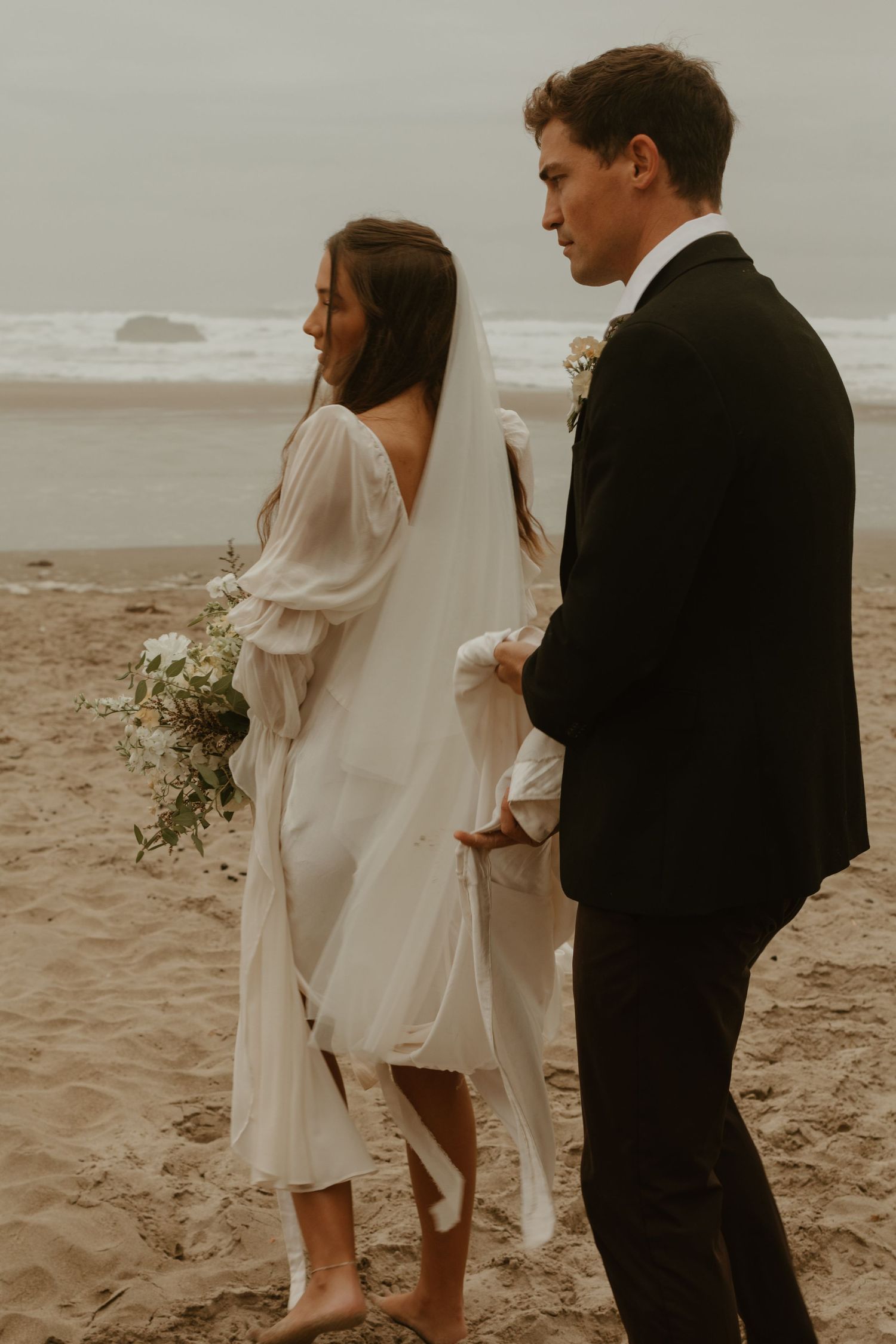 Bride in white dress with veil and groom in black suit stand barefoot on a misty beach.