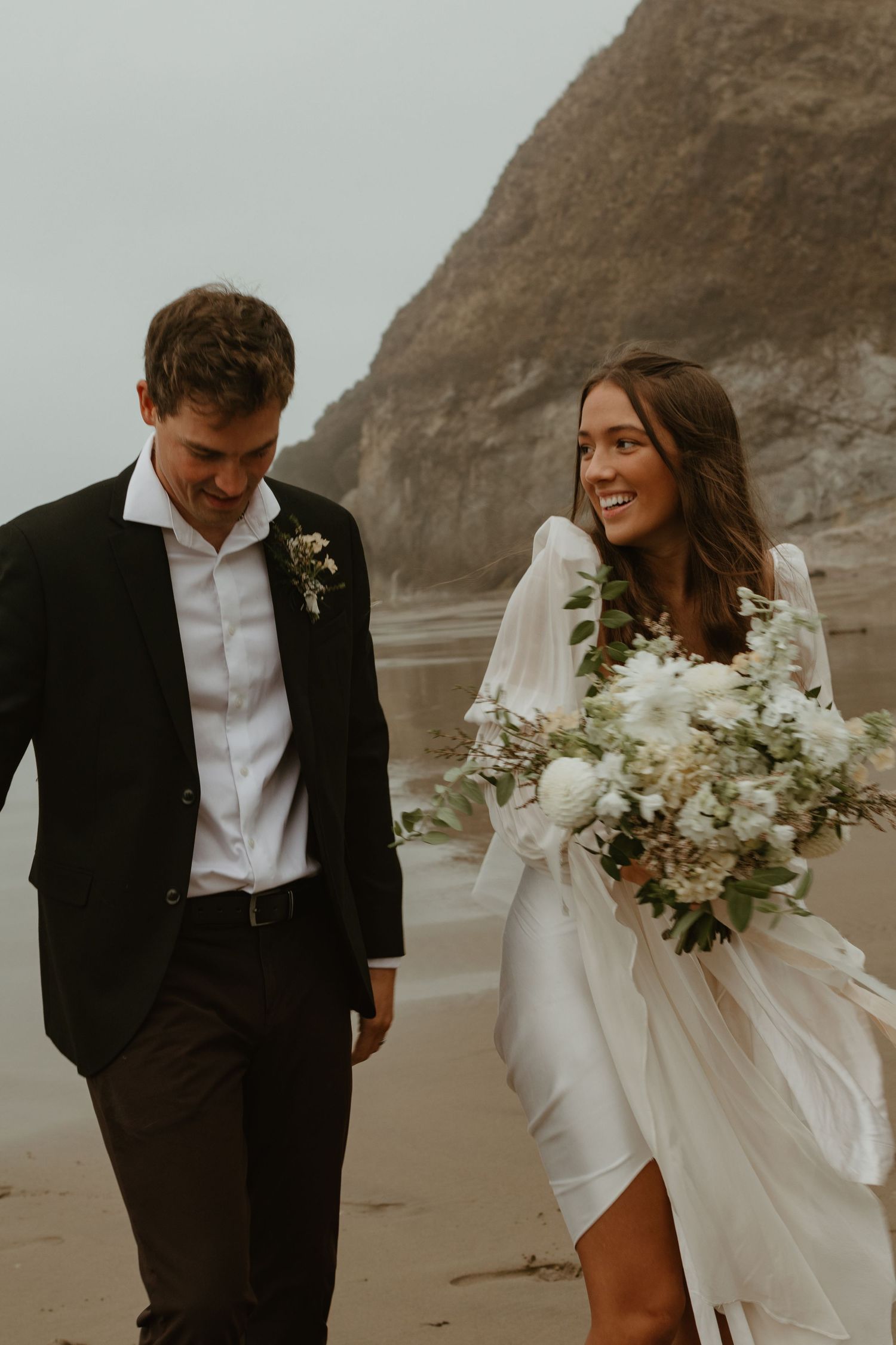 Smiling bride holding white floral bouquet walks with groom along a rocky shoreline.