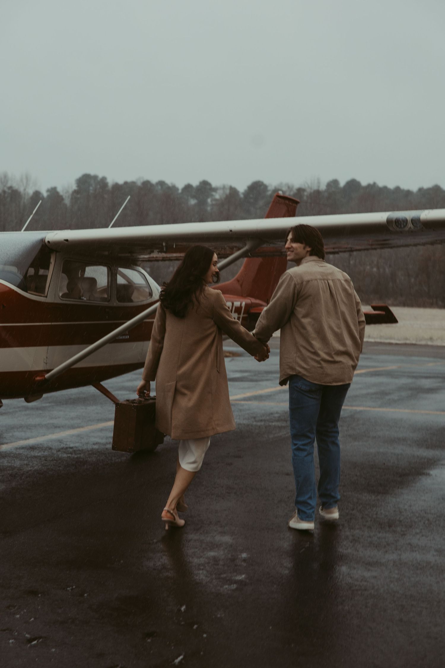 Couple holding hands walking toward a vintage red airplane on a wet rainy airstrip.