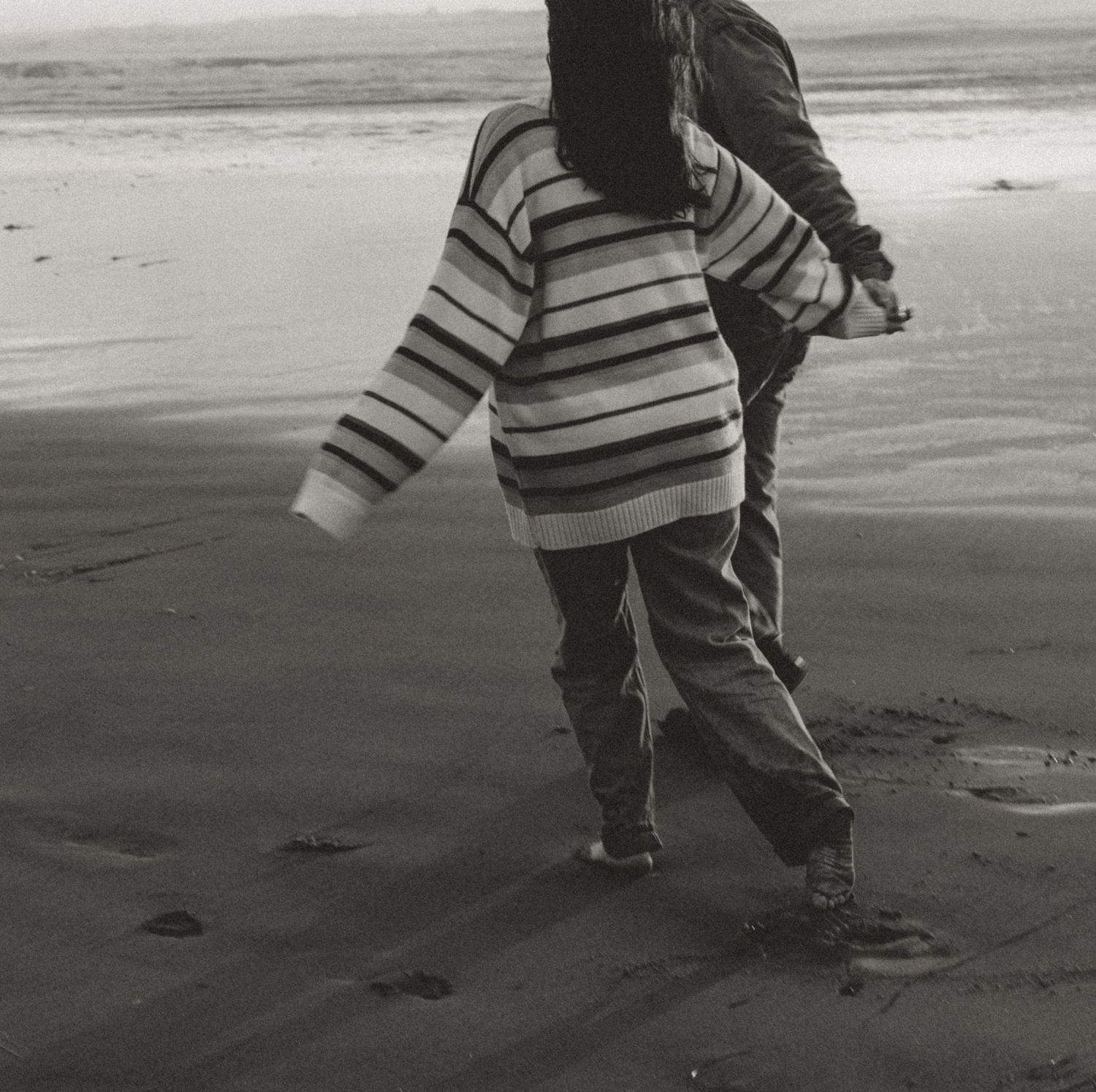 Black and white photo of couple walking barefoot together along a windy ocean beach.