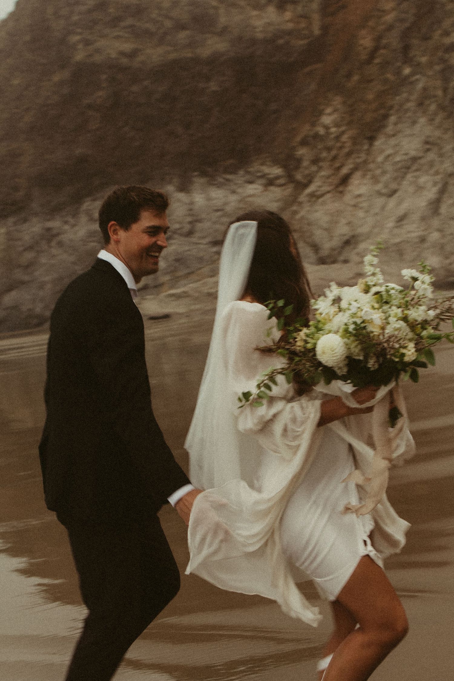Bride in white dress with veil and bouquet running with groom on a dramatic coastal beach with rocky cliffs.