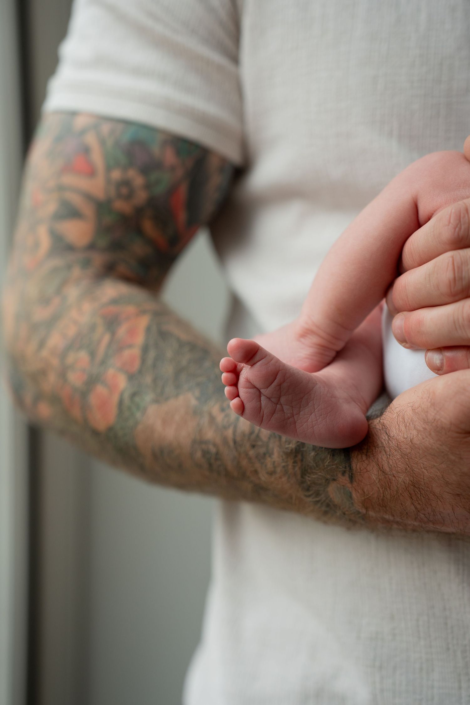 Tattooed father's arm gently holding tiny newborn baby feet against white shirt.