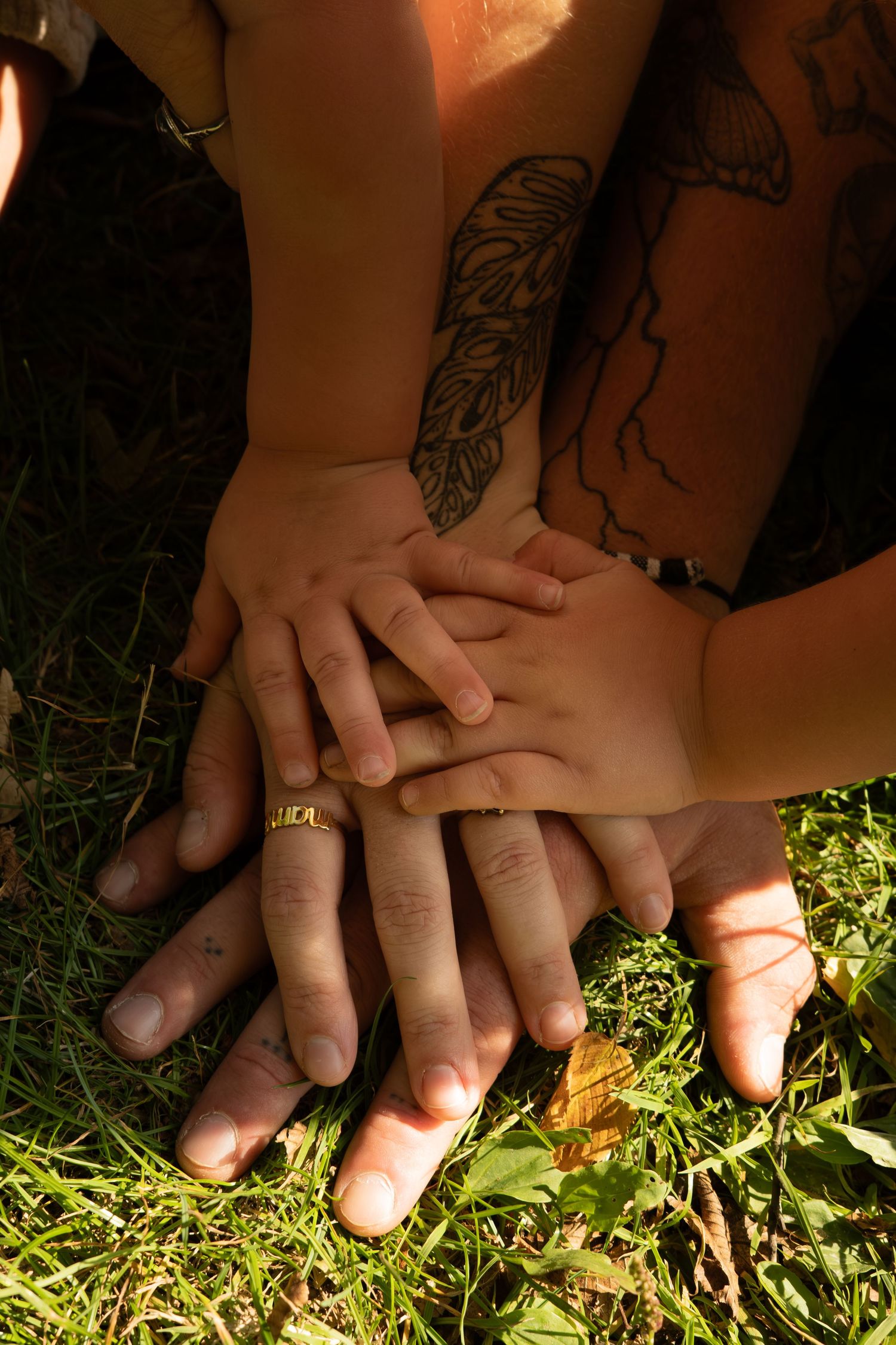 Family hands stacked together on green grass, showing tattoos and a gold name ring.