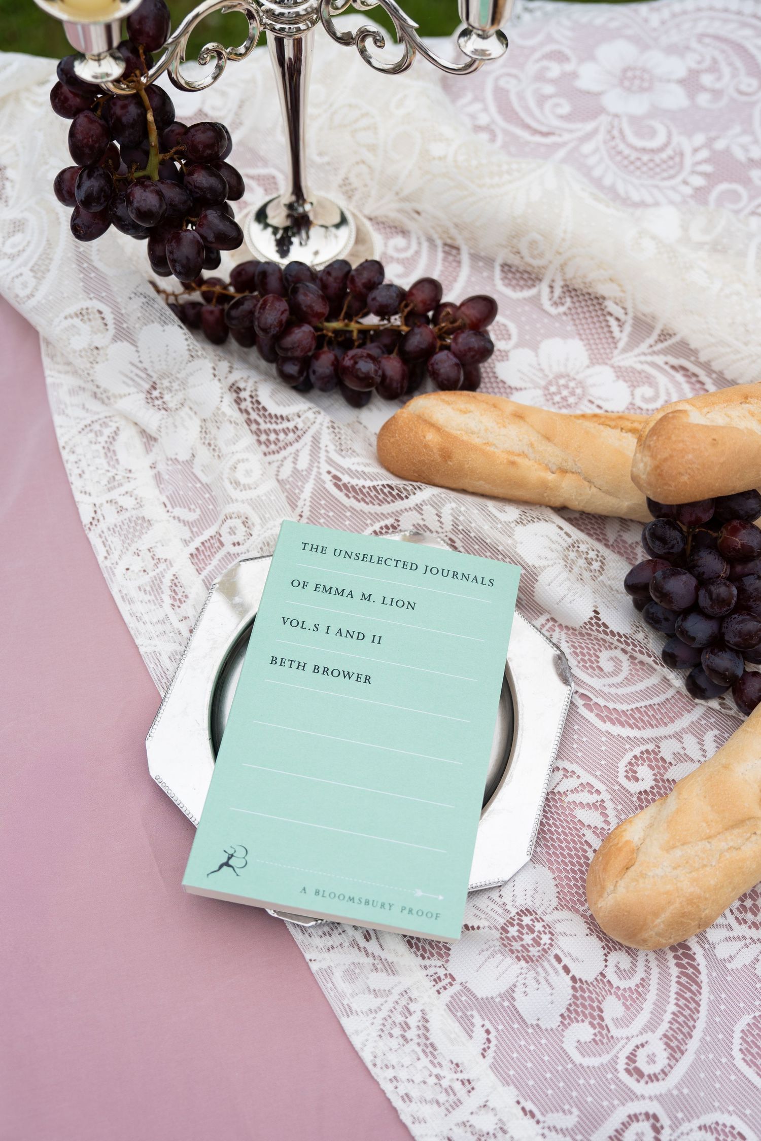 Elegant table setting with mint book, purple grapes, bread, lace cloth, and silver candlestick on pink background.