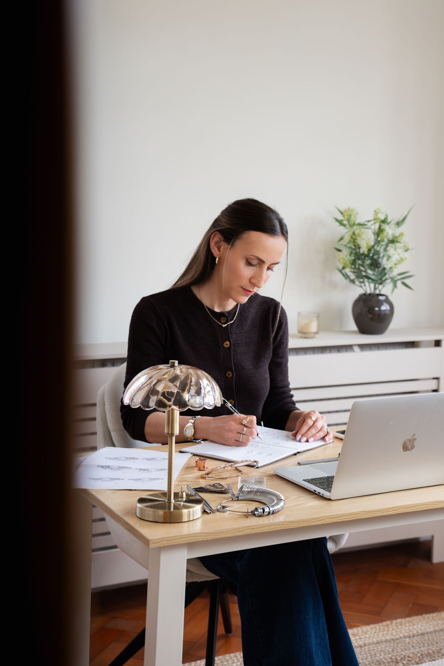 Professional woman in black cardigan writing notes at wooden desk with laptop and gold lamp in bright home office.
