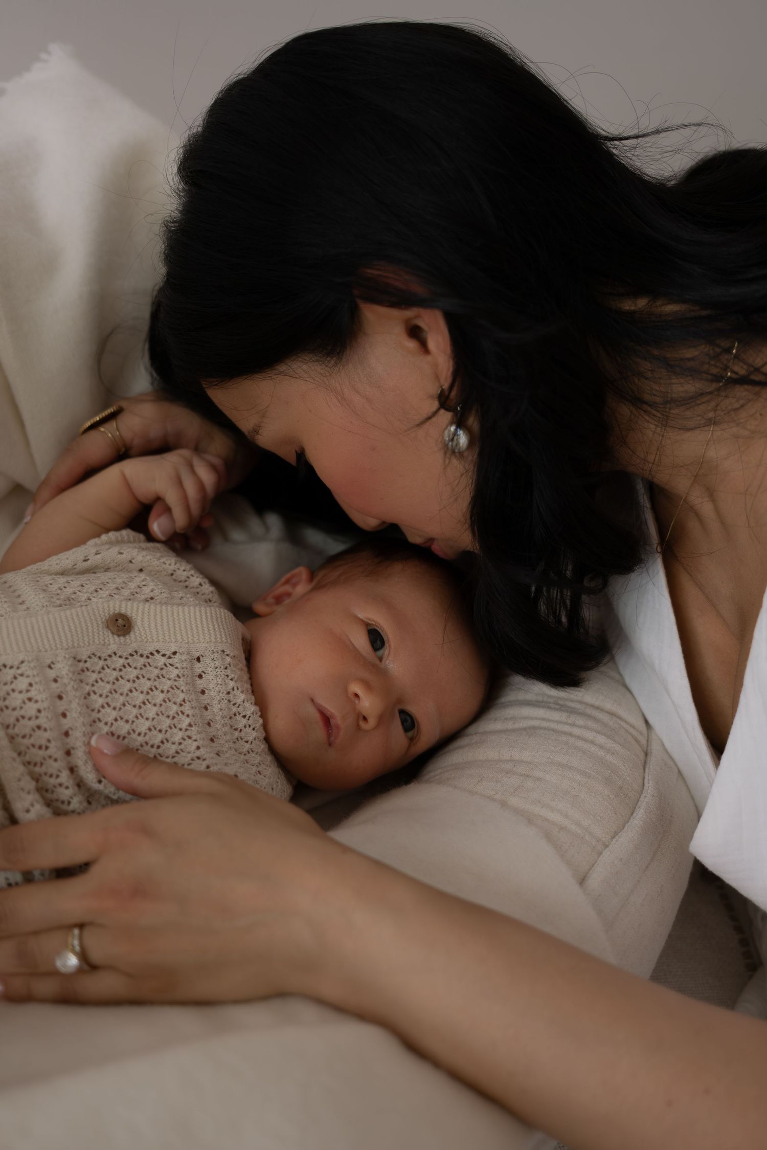 Tender moment of dark-haired mother kissing her newborn baby wrapped in a knit blanket.