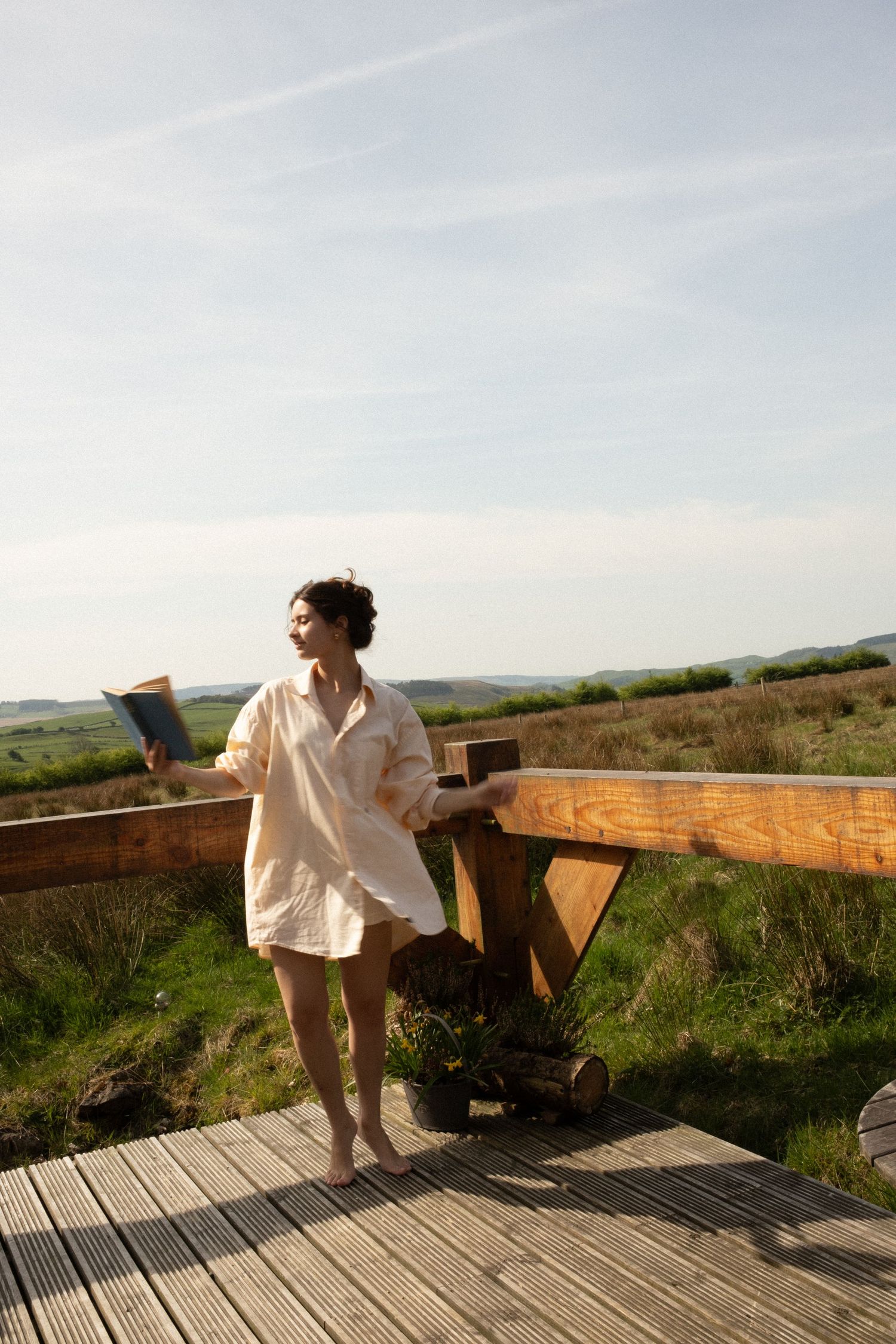 Woman in white linen shirt holding a book on a wooden deck overlooking a scenic green countryside landscape.