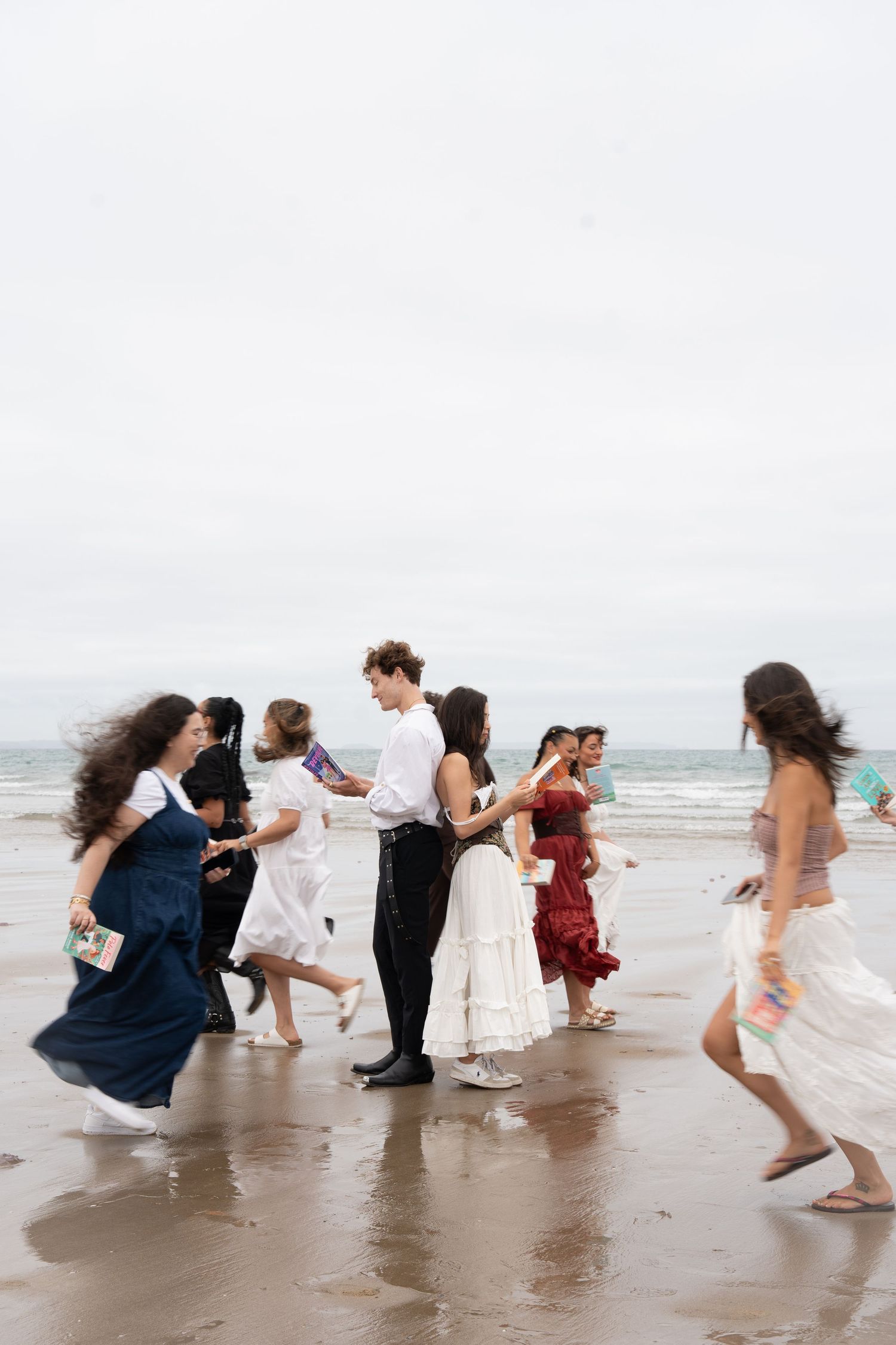 Group of people in elegant dresses dancing and running on a sandy beach on an overcast day.