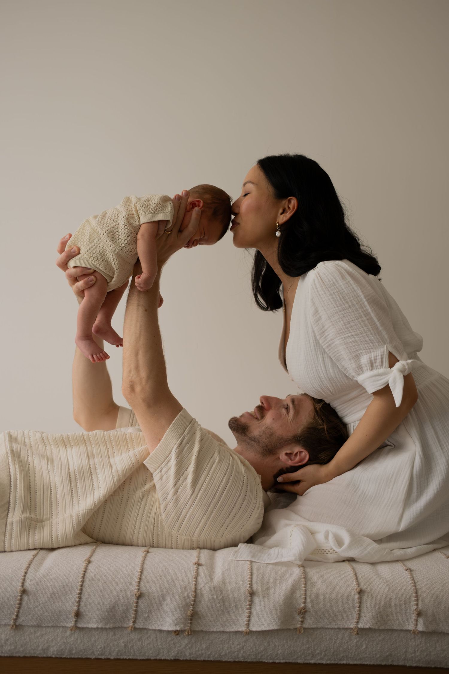 New parents tenderly kiss their newborn baby while relaxing together on a white bed at home.