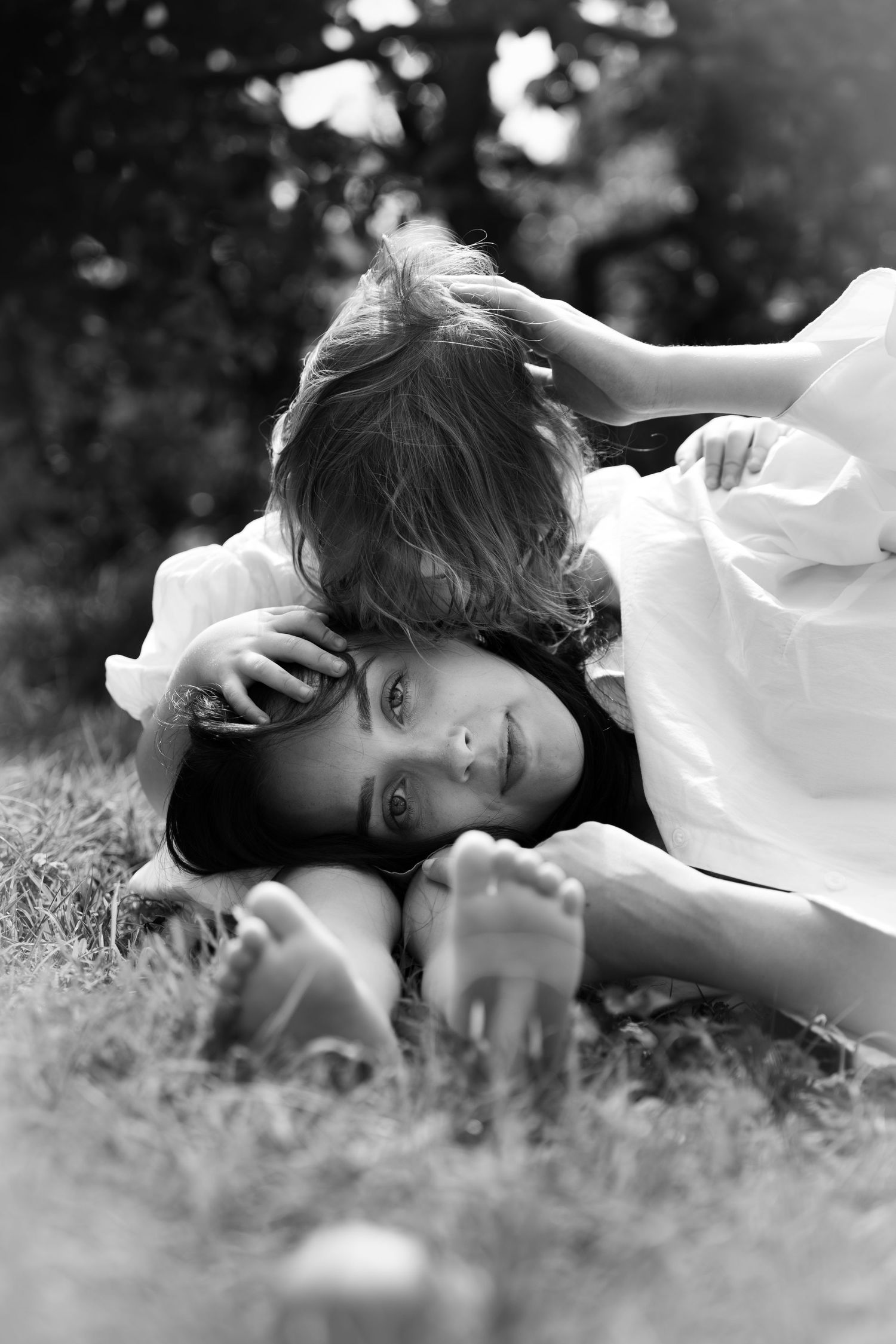 Black and white photo of mother lying on grass while young child playfully hugs her head.