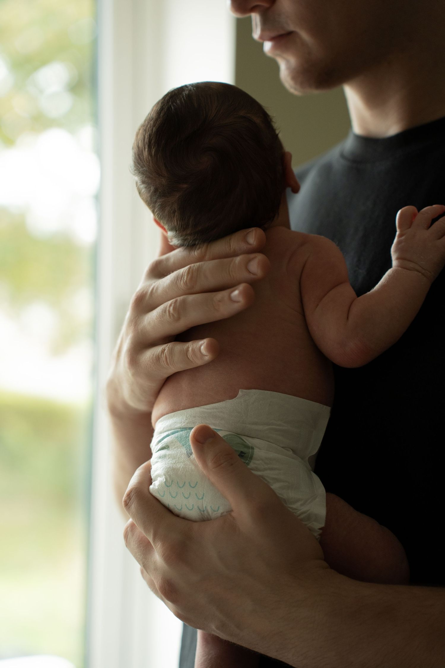 Father in dark shirt holding newborn baby in diaper against bright window with green view.