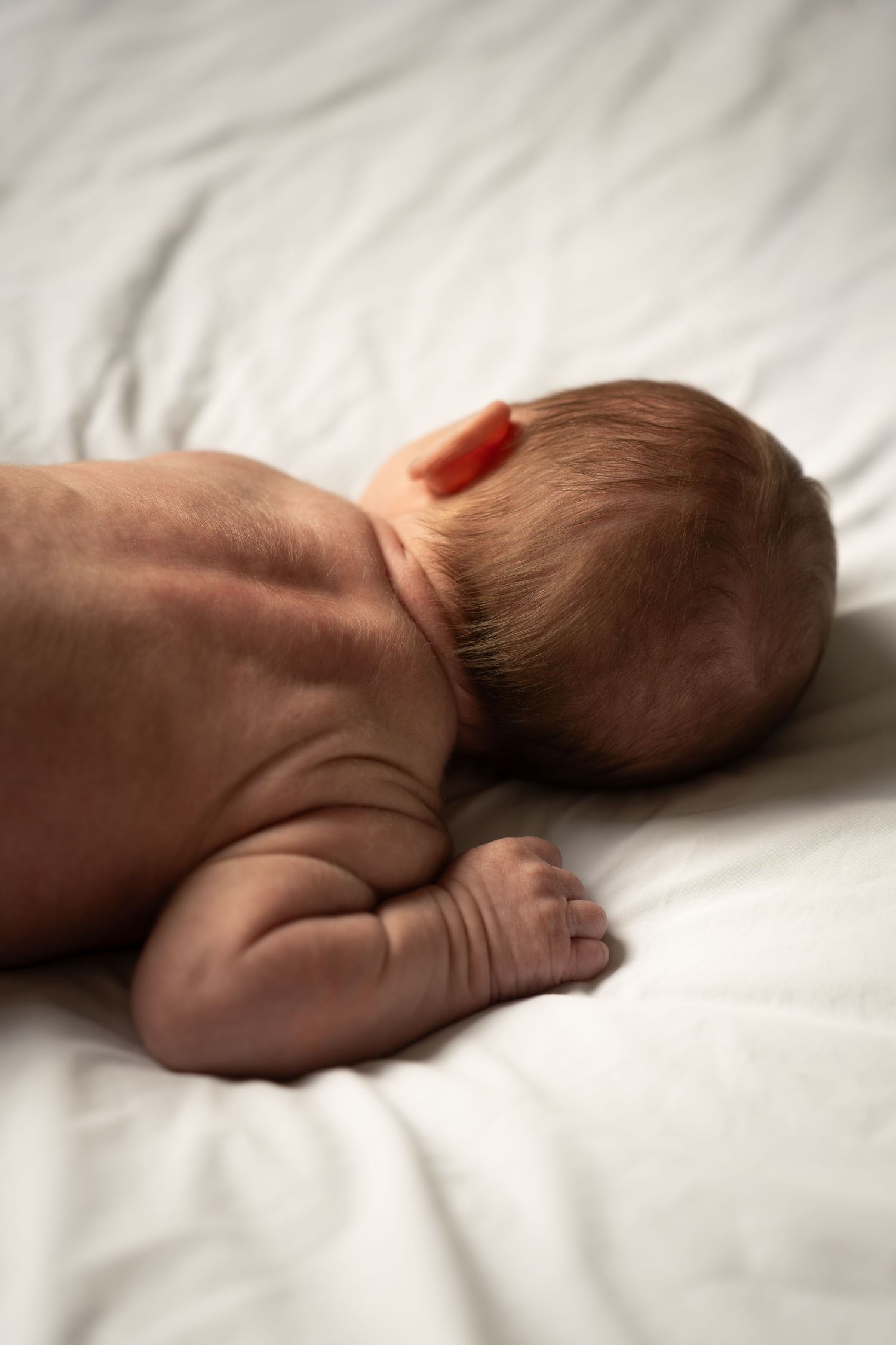 Newborn baby lying on white bedding, photographed from behind showing tiny feet and back.