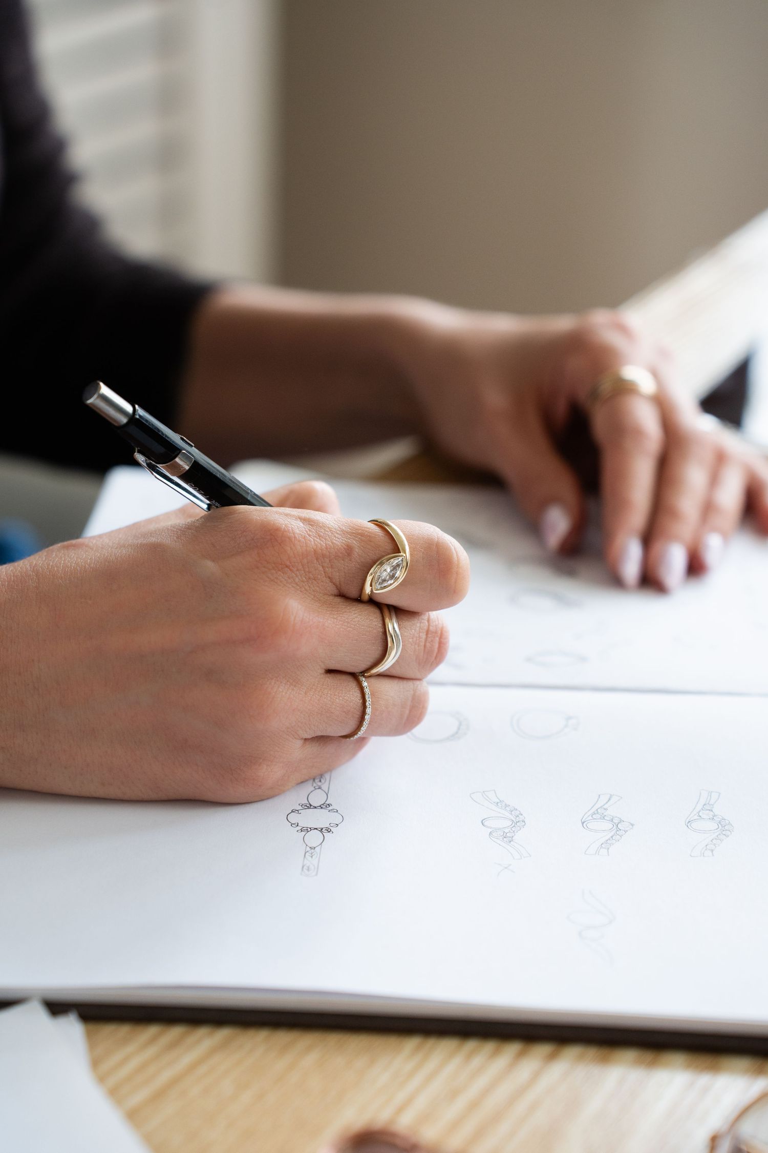 Close-up of hands wearing gold rings sketching jewelry designs on white paper at a wooden desk.