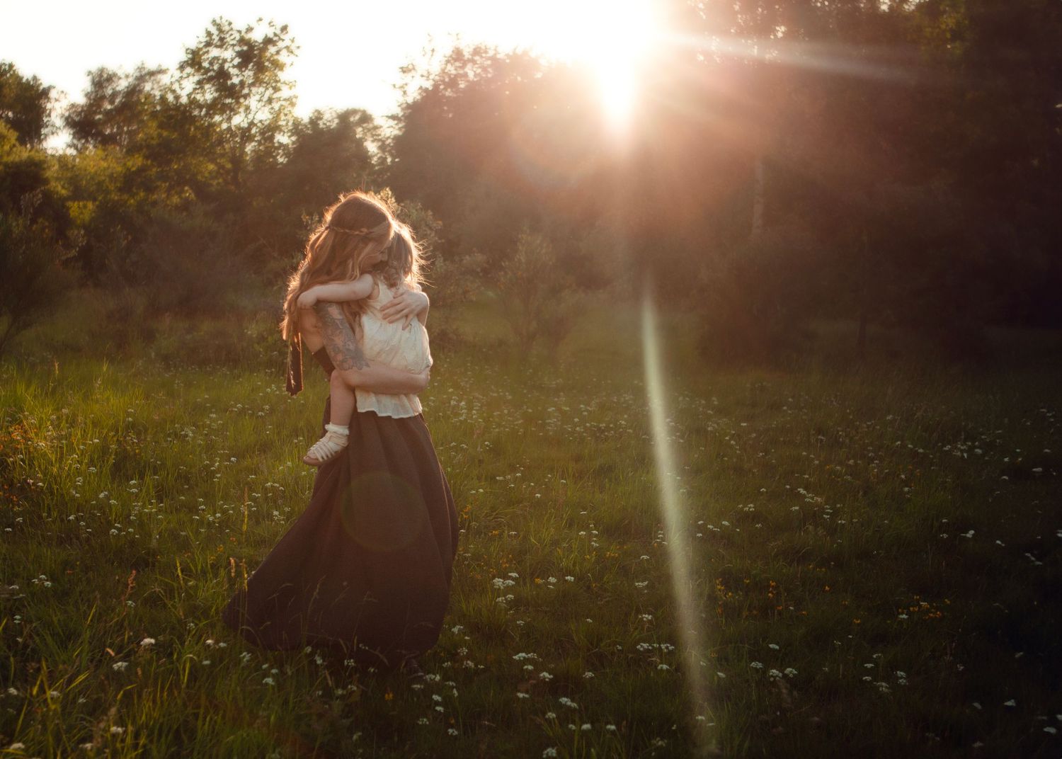 Woman in dark skirt holds child in sunlit meadow with trees, golden hour light creating lens flare.
