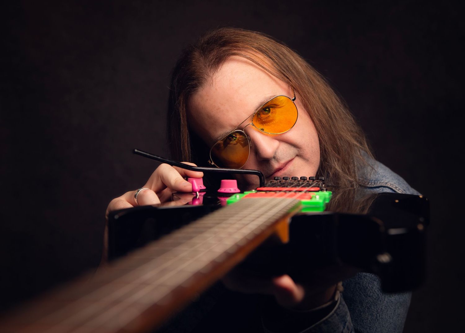 A musician with long hair and orange tinted glasses examines an electric guitar neck up close, in Kidderminster