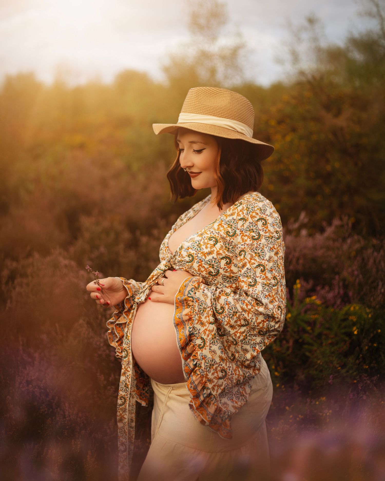 Pregnant woman in floral kimono and straw hat cradling baby bump in golden hour field of purple heather in the West Midlands