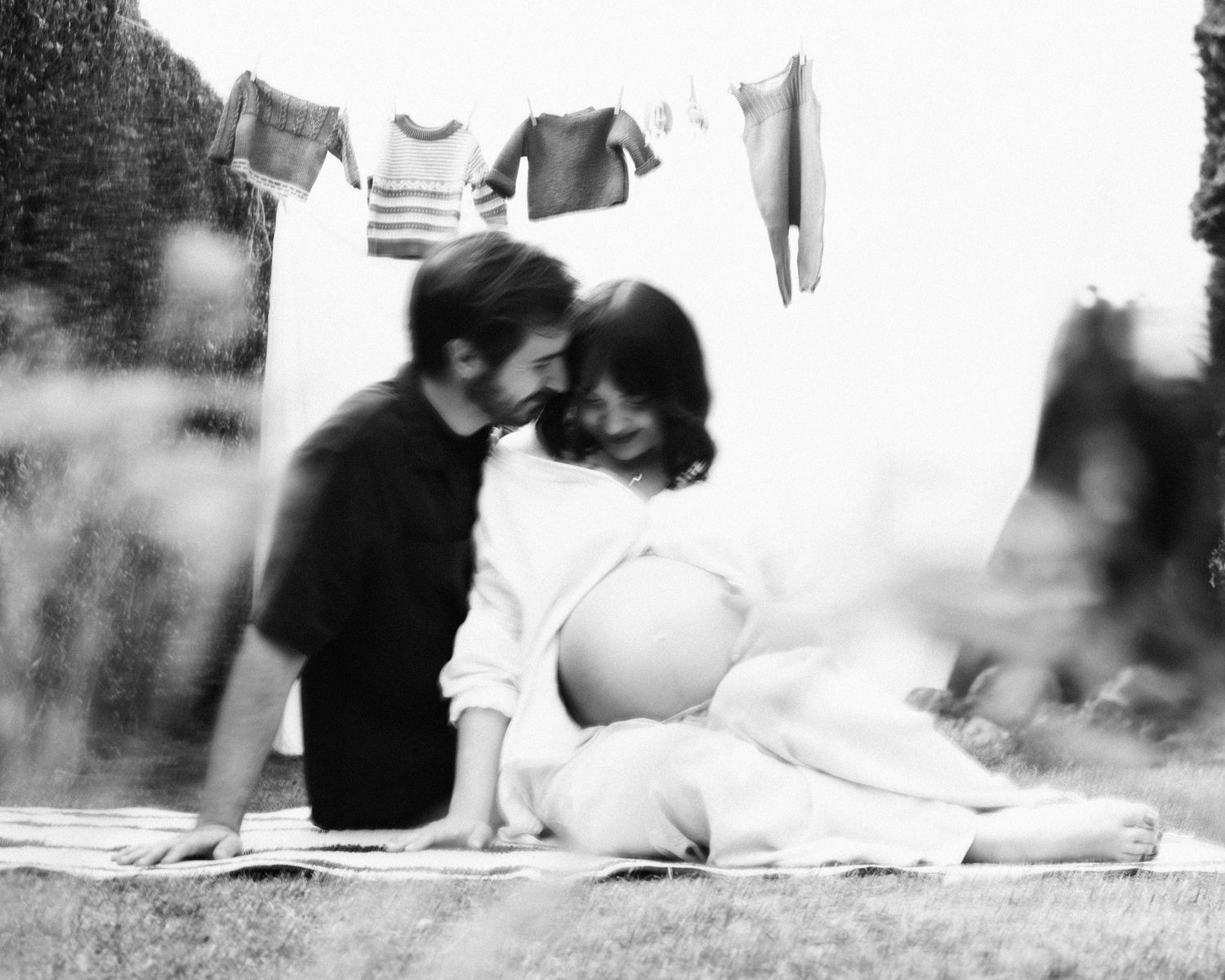 Black and white photo of couple sitting on grass under clothesline with baby clothes hanging  in Birmingham