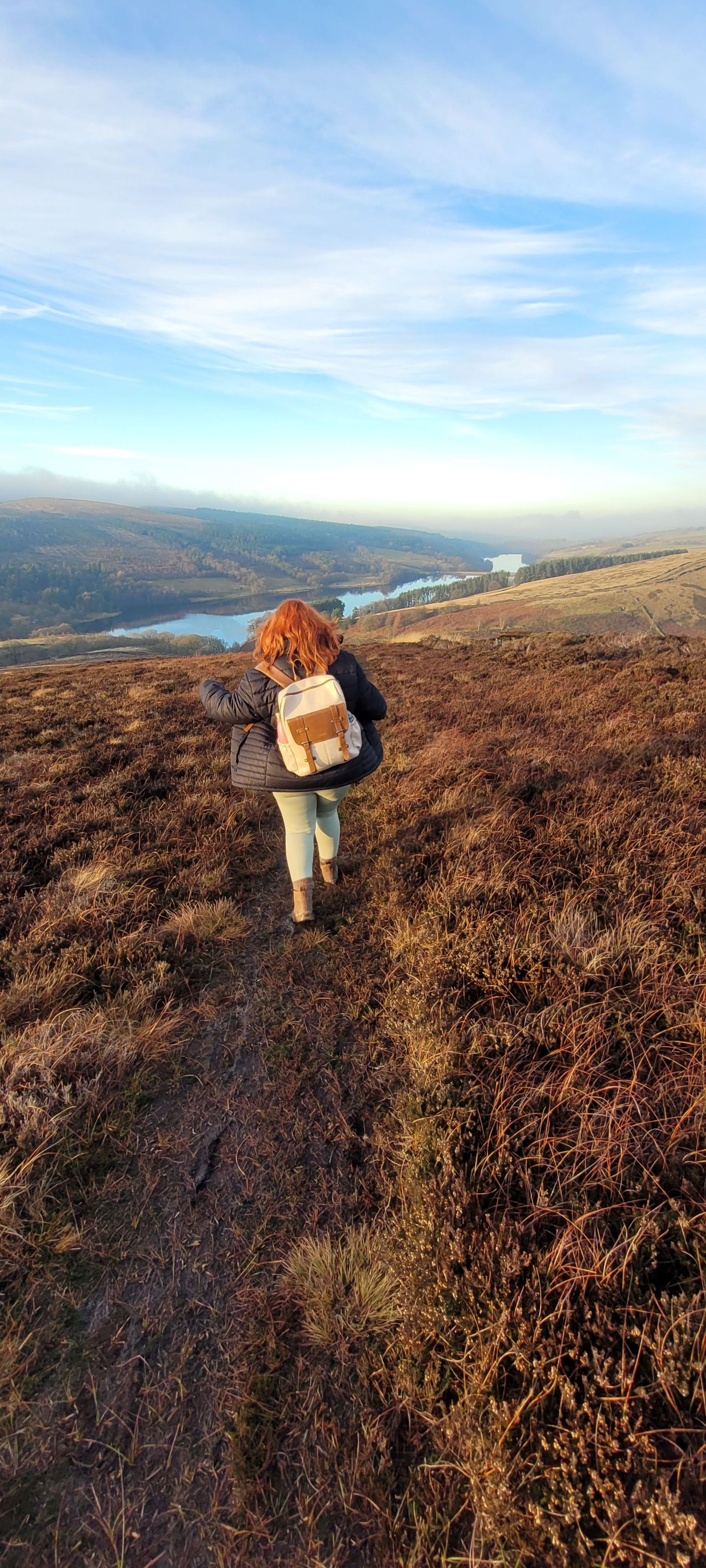 Redhead woman with backpack hiking along a moorland trail with a scenic reservoir and blue sky behind her in West Midlands