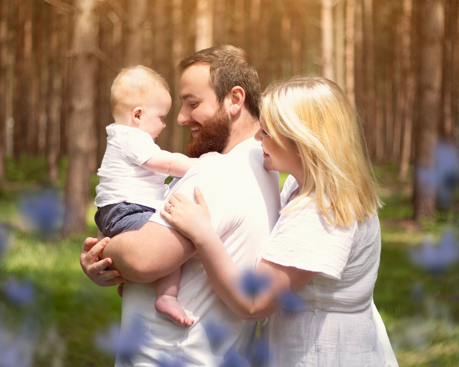 A young family with a bearded father holding a baby while a blonde mother looks on in a sunlit forest in the West Midlands