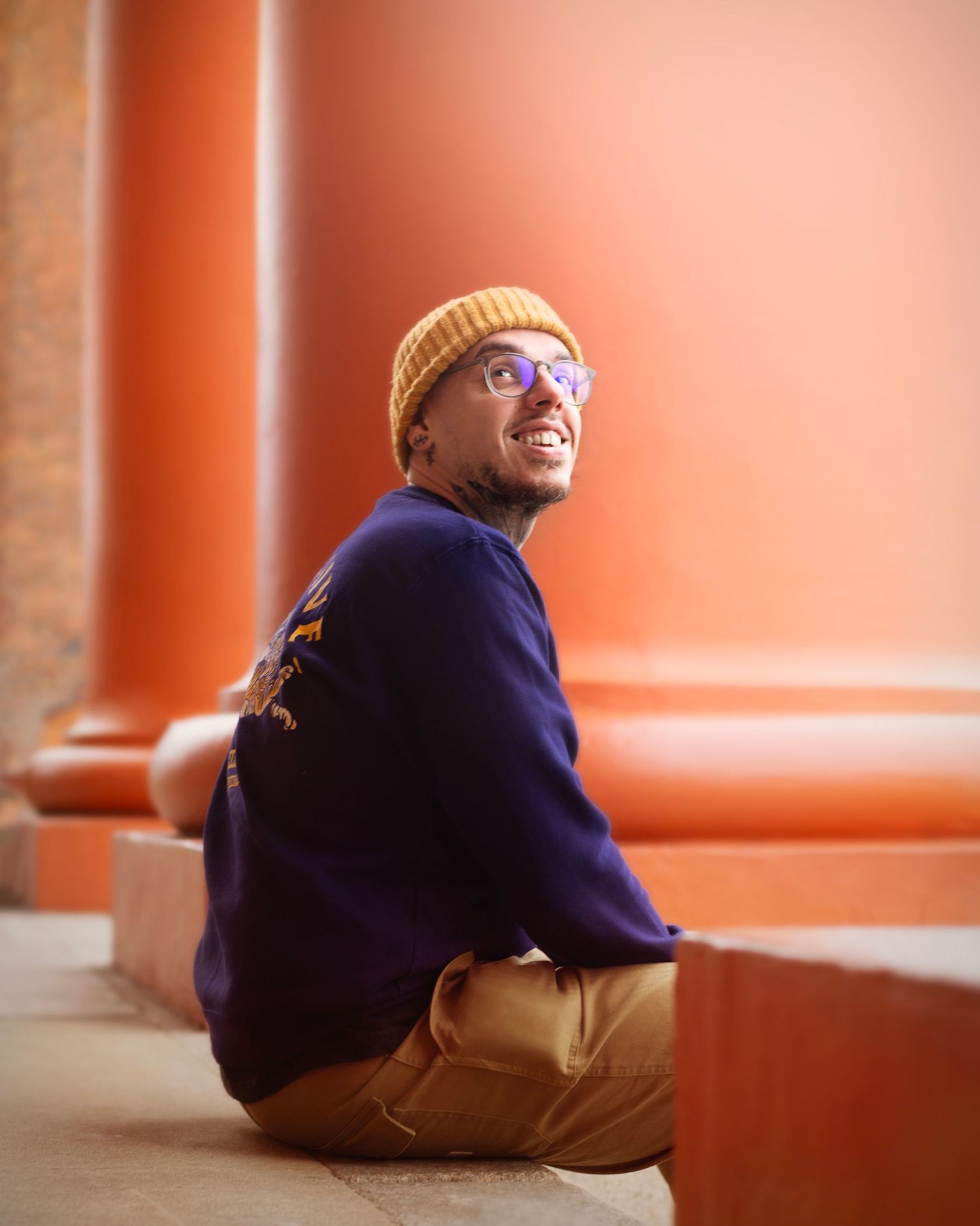 A smiling young Black man in a yellow beanie and navy sweater sits against an orange wall outdoors in the West Midlands