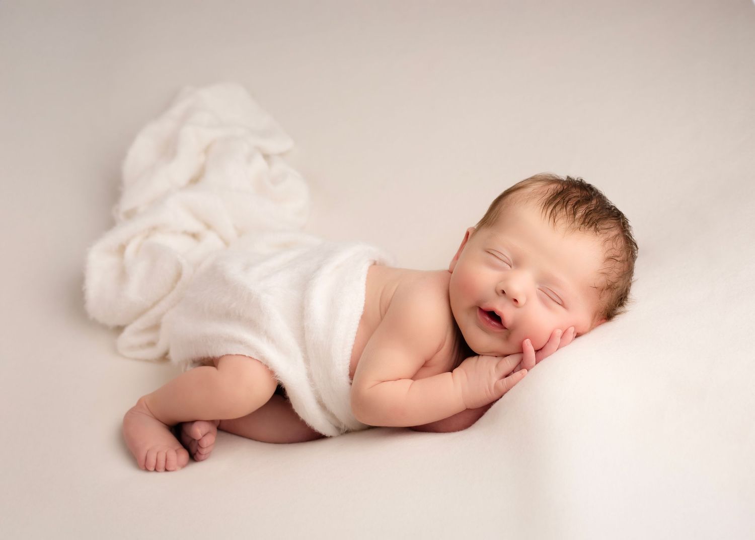 Smiling newborn baby wrapped in white blanket lying on soft white background  in Birmingham