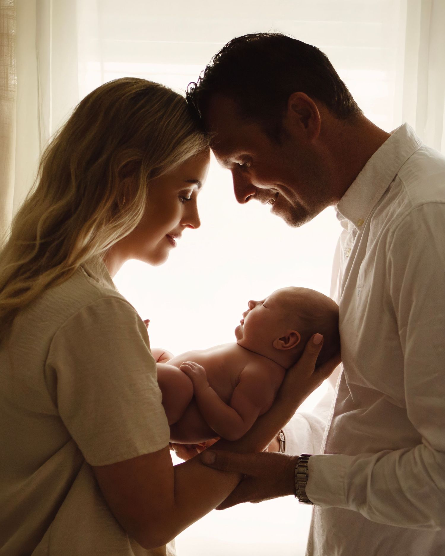 A couple smiles tenderly at their newborn baby held between them, backlit by soft window light.