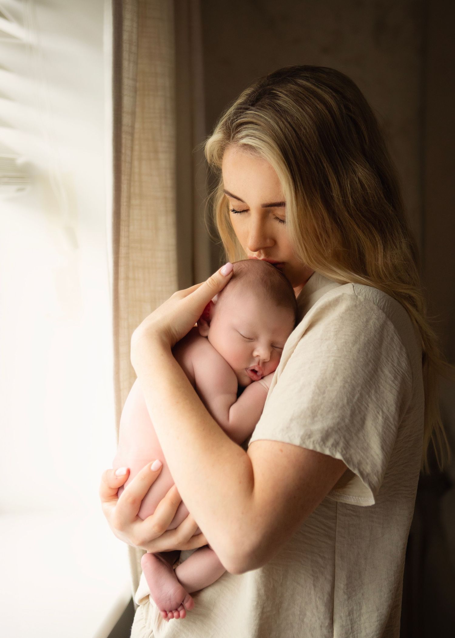A tender mother gently kisses her sleeping newborn baby while standing by a softly lit window.