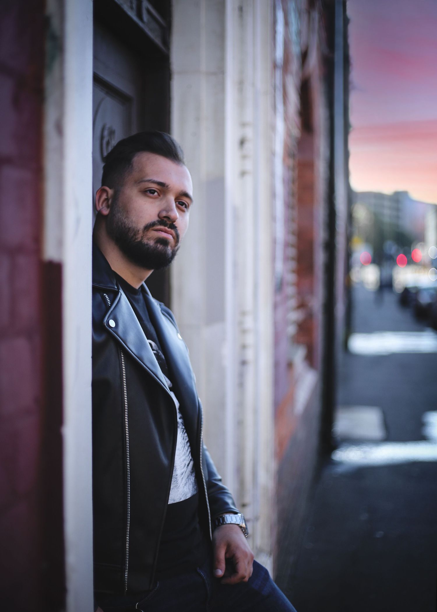 Bearded man in black leather jacket leaning against building wall at twilight on city street in the West Midlands