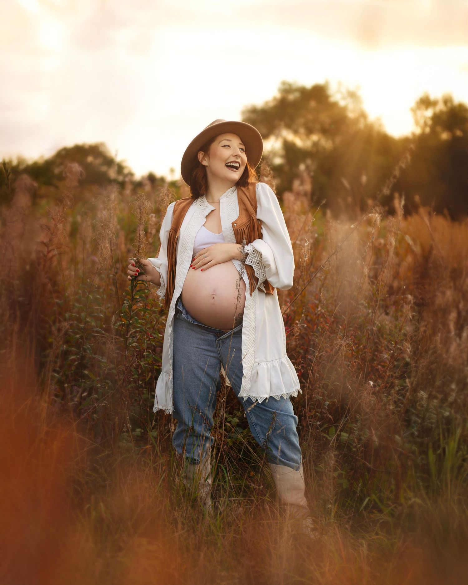 Joyful redhead pregnant woman in hat and jeans laughing in a golden sunset wildflower field in the West Midlands