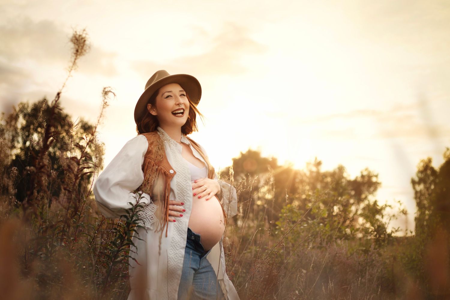 Happy pregnant Asian woman in hat and white kimono laughs while holding belly in sunset field  in Birmingham