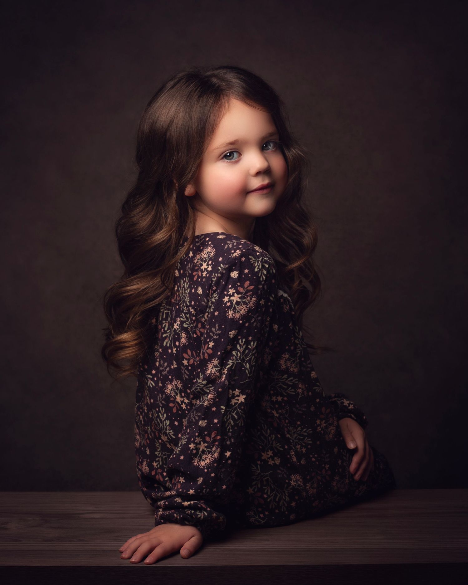Young girl with long wavy brown hair poses in a floral dark dress against a moody studio background in the West Midlands