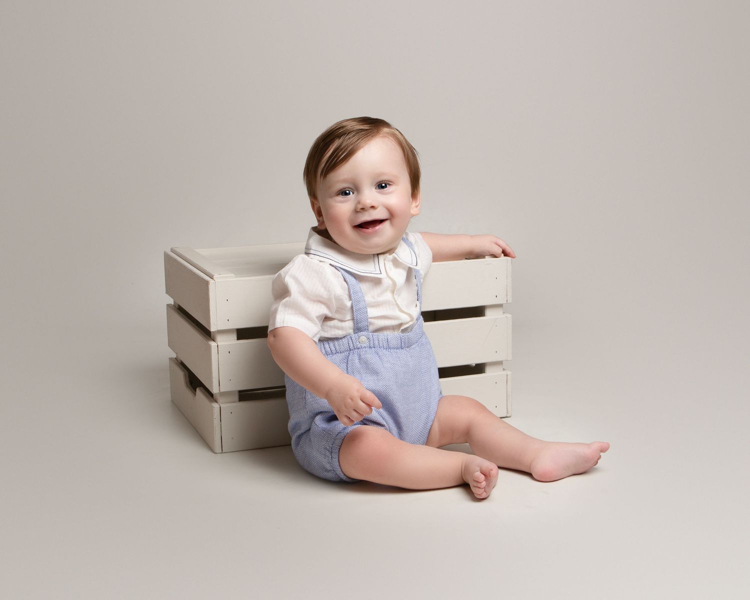 Smiling baby boy in blue overalls and white shirt sits beside a wooden crate in a studio portrait in the West Midlands