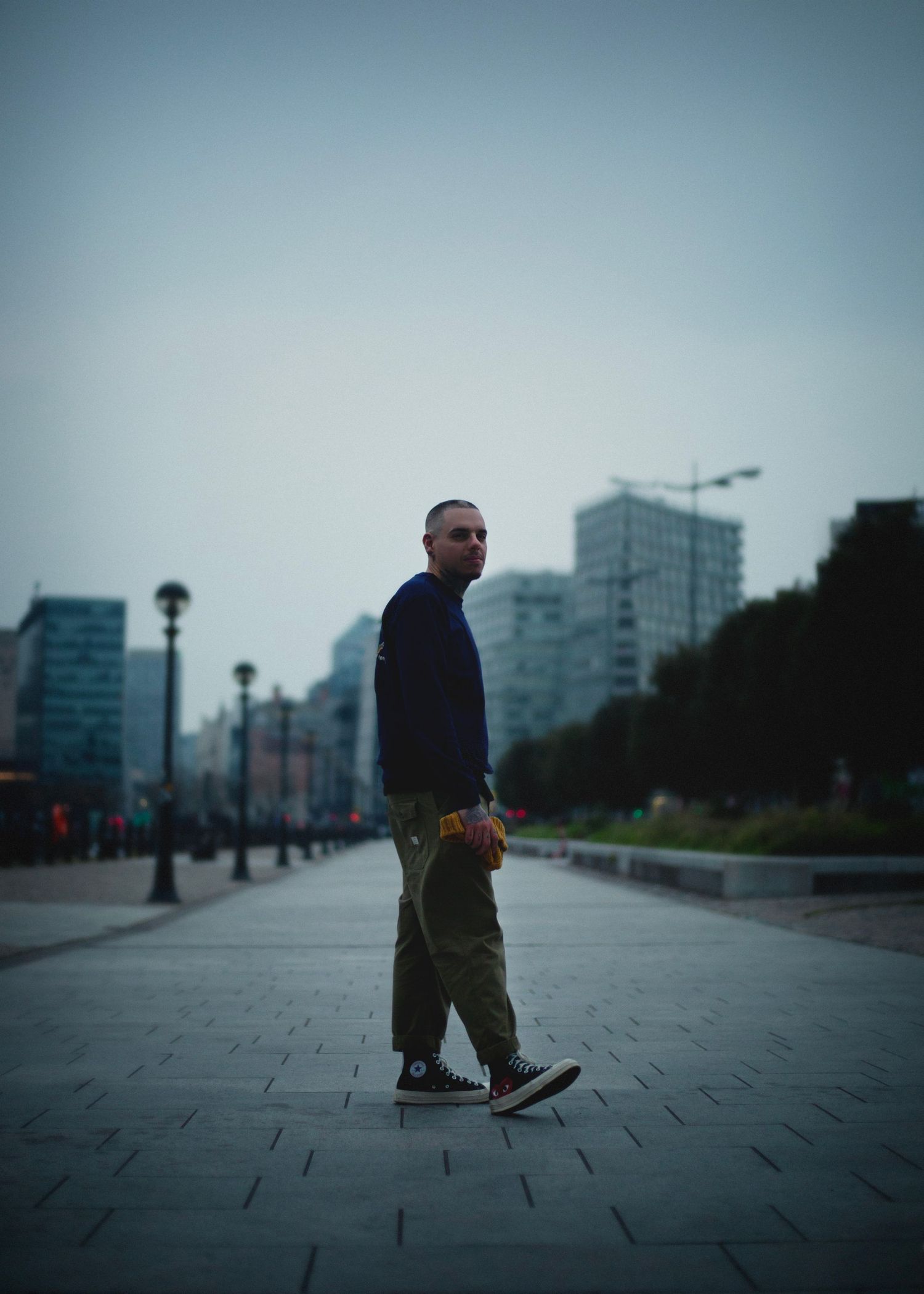 A bald man in a navy sweatshirt and cargo pants walks along an urban plaza at dusk with city buildings in Birmingham